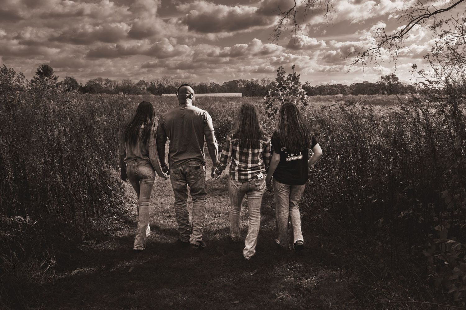 Four friends walk hand-in-hand down a rural path through tall grass under dramatic cloudy skies in sepia tone.