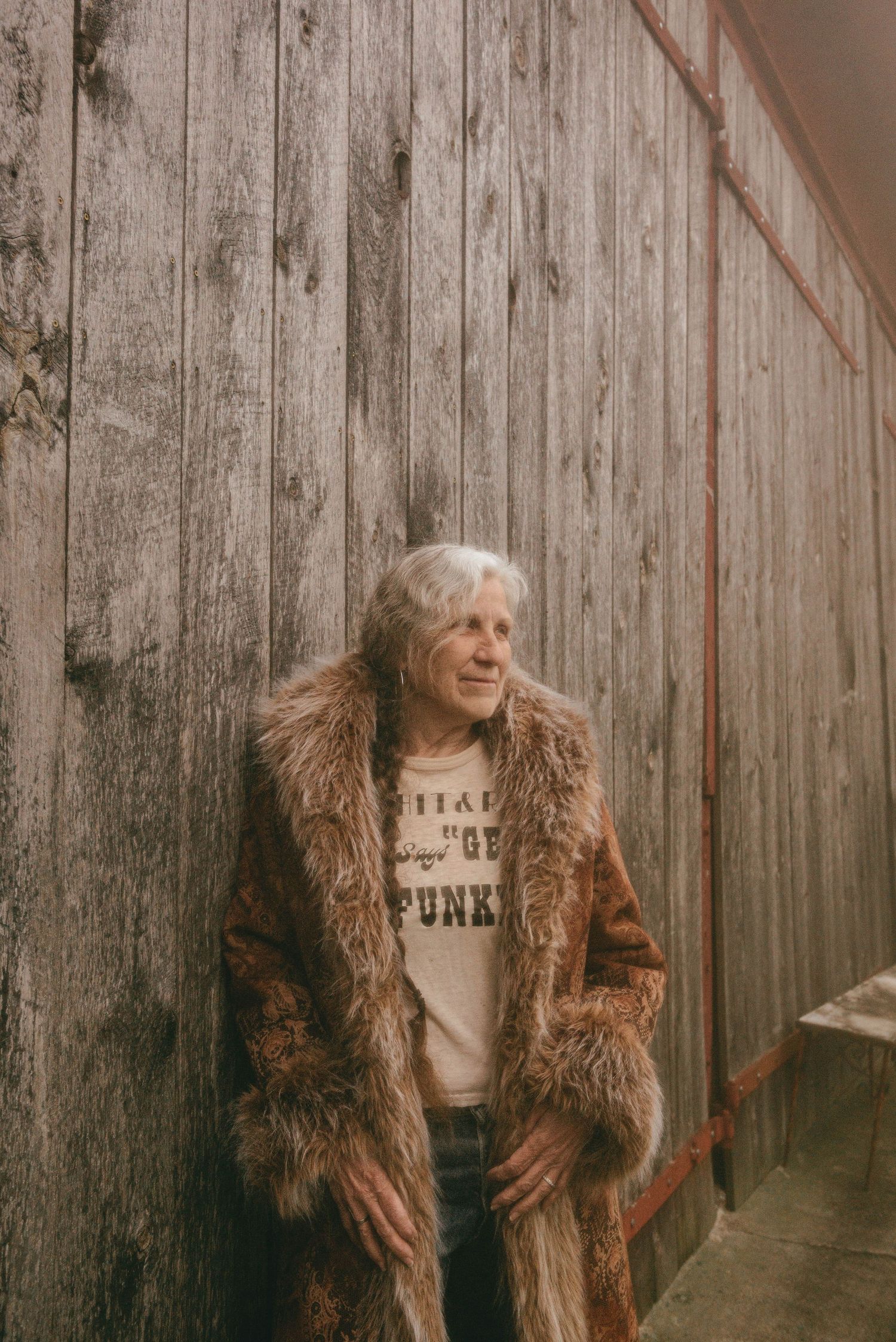 Elderly woman with white hair wearing a fur coat leaning against a rustic wooden wall.