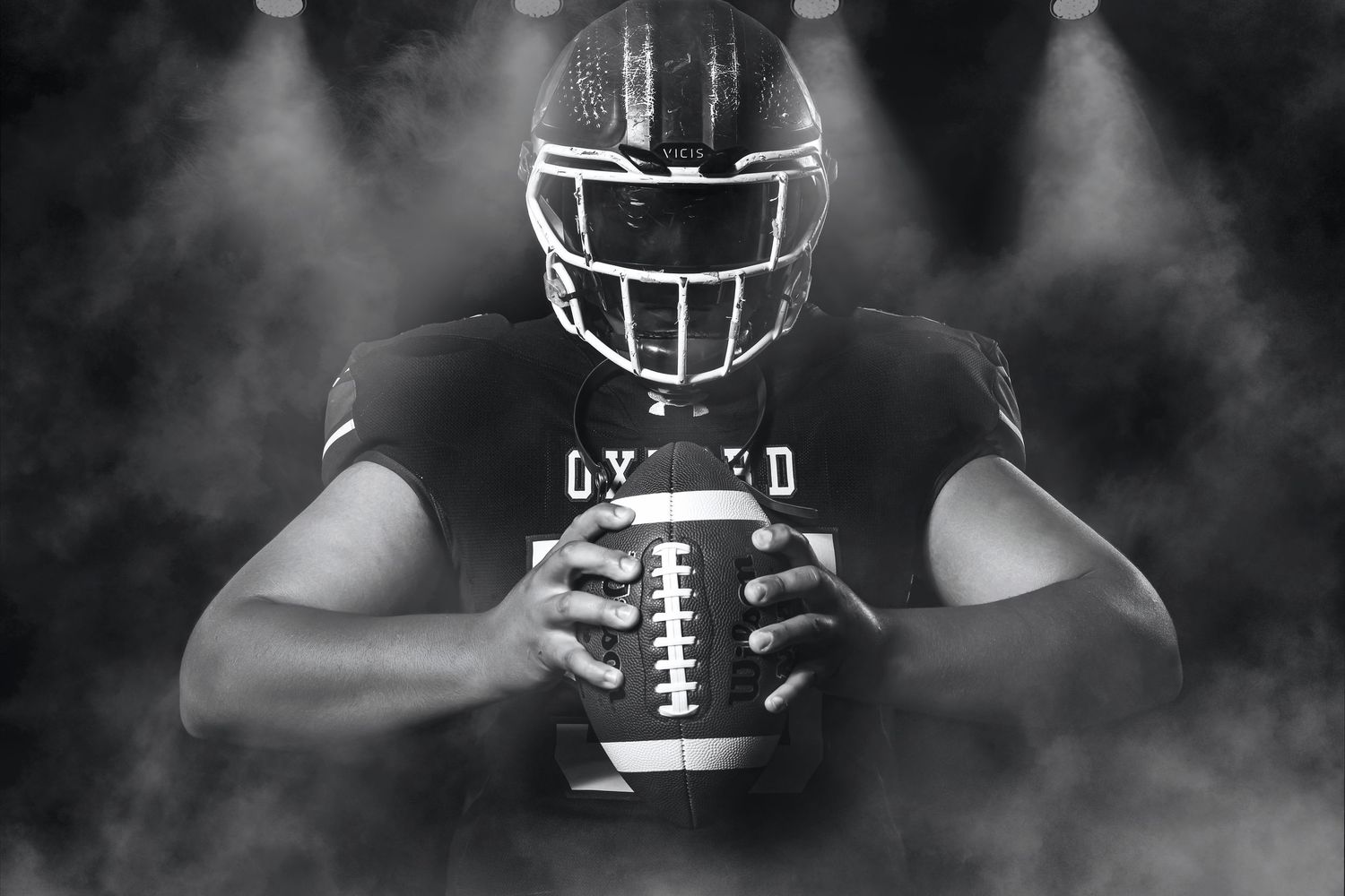 Black and white football player in Oxford jersey holding ball in smoky stadium.