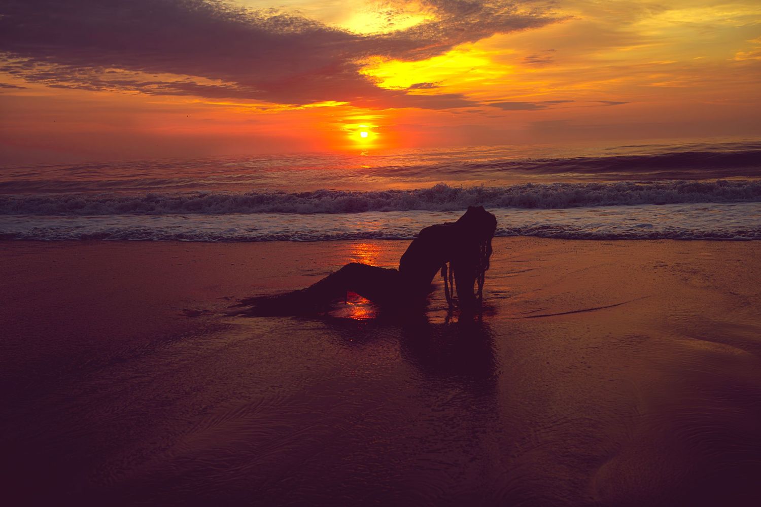 Silhouette of a person on a sandy beach at golden sunset with waves and vibrant orange sky.