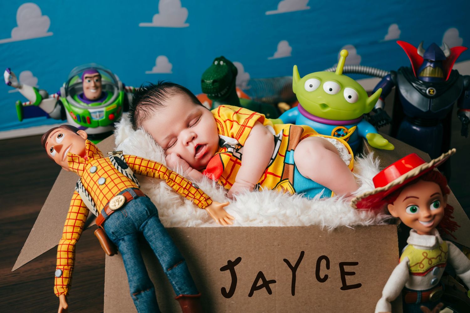 Newborn baby named Jayce sleeps in a box surrounded by Toy Story character dolls including Woody, Buzz, and Jessie.