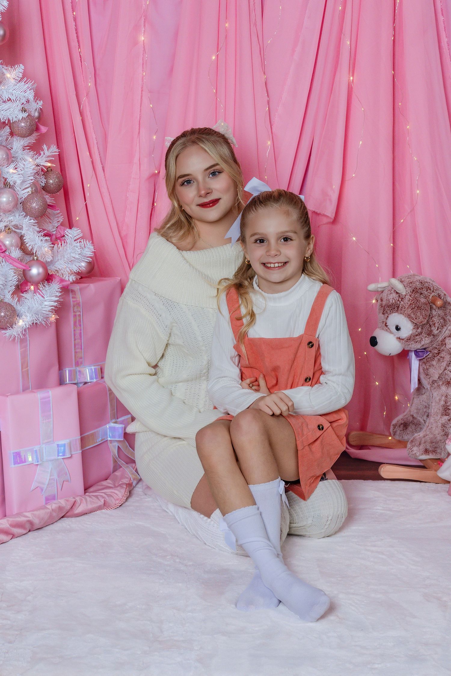 Two smiling girls pose together in a pink Christmas-themed room with gifts, a white tree, and a teddy bear.