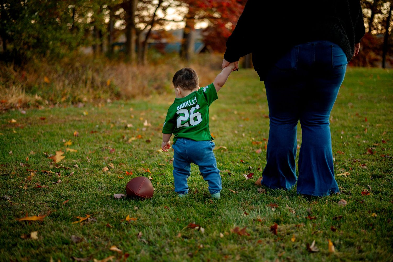 Toddler in green jersey #26 holds adult's hand on autumn grass with football nearby.