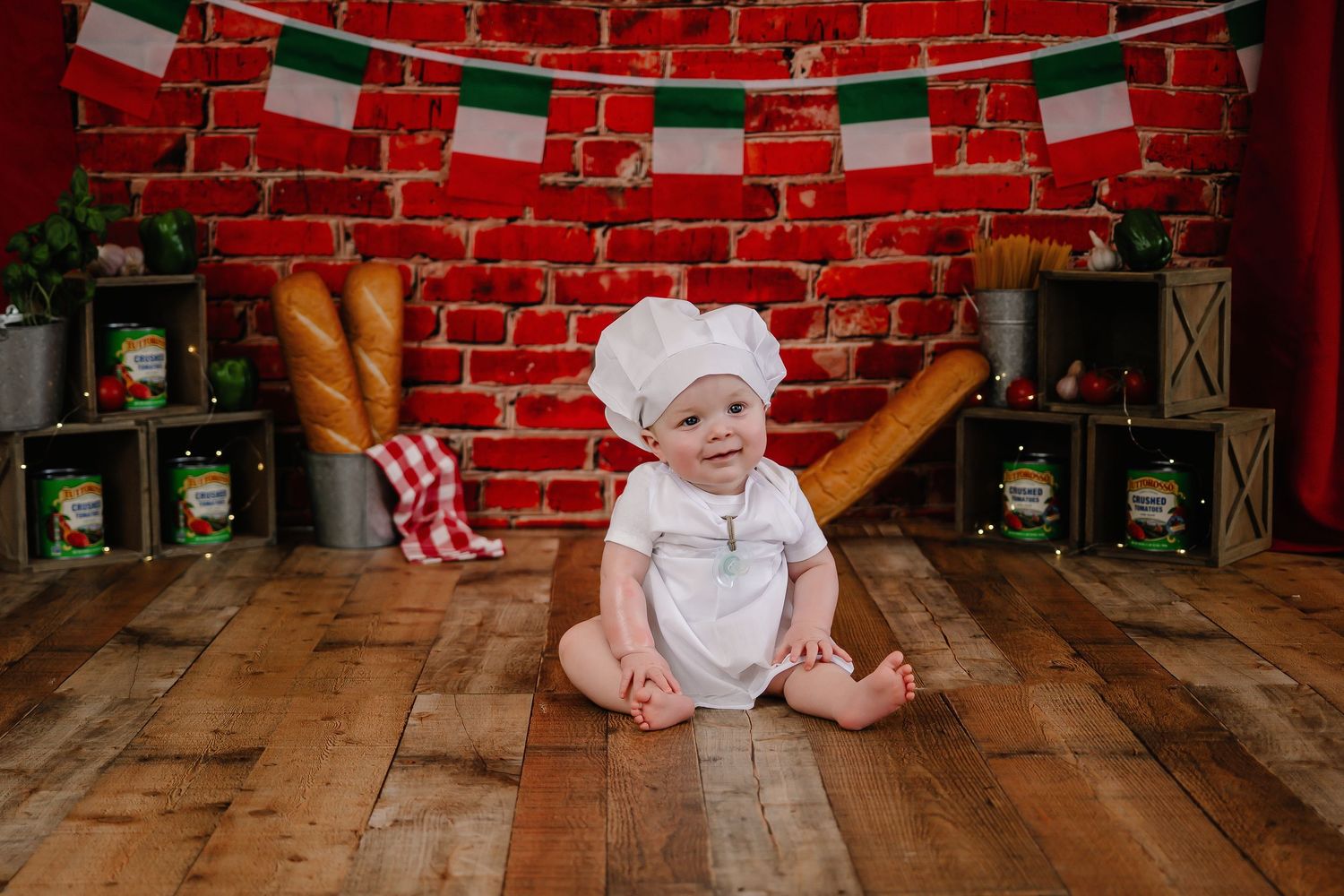 Adorable baby in chef hat and white outfit sitting on wood floor with Italian-themed backdrop.