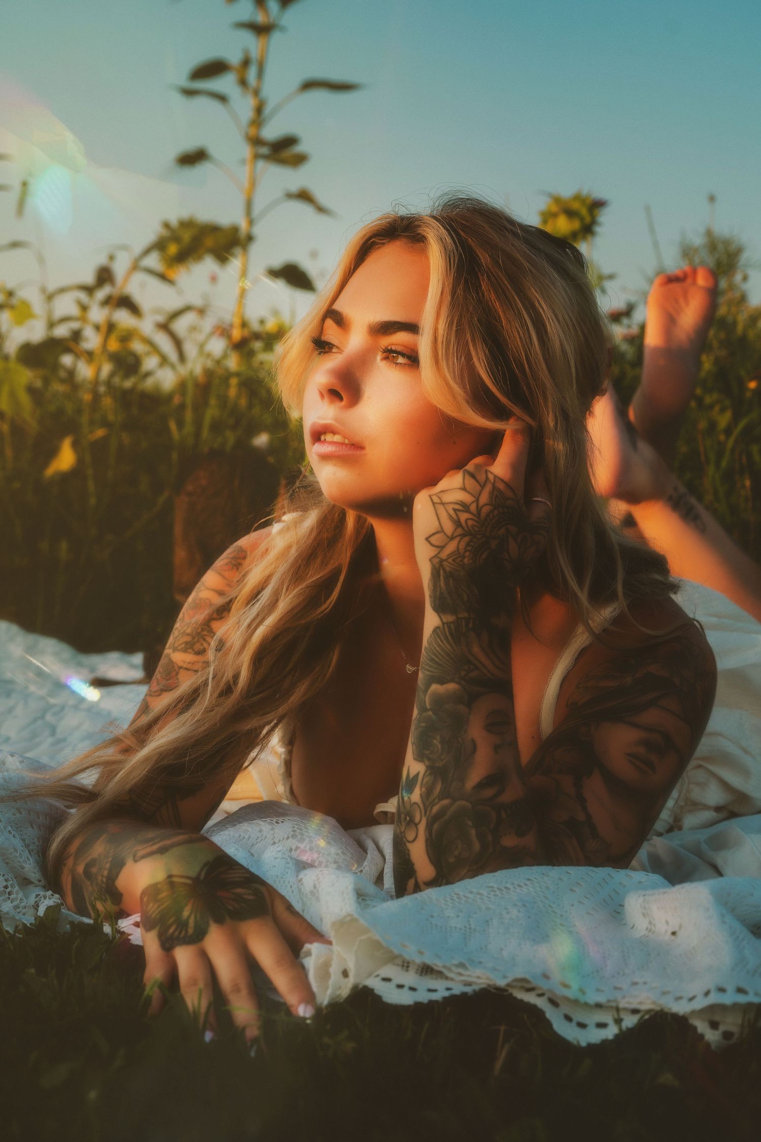 Tattooed blonde woman lying on a blanket in a sunlit field, gazing thoughtfully with golden hour light.