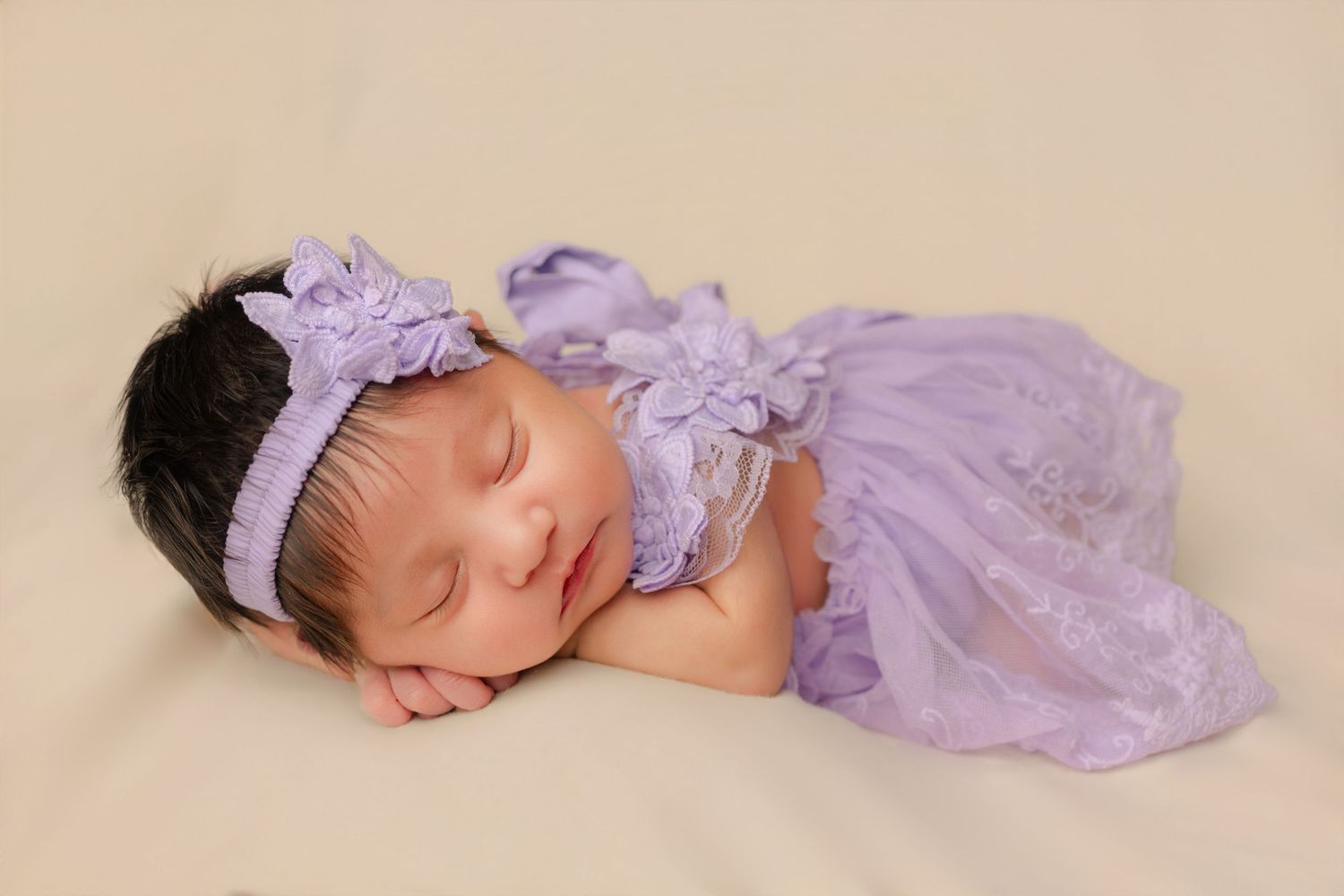 Sleeping newborn baby girl in purple lace outfit and floral headband on cream background.