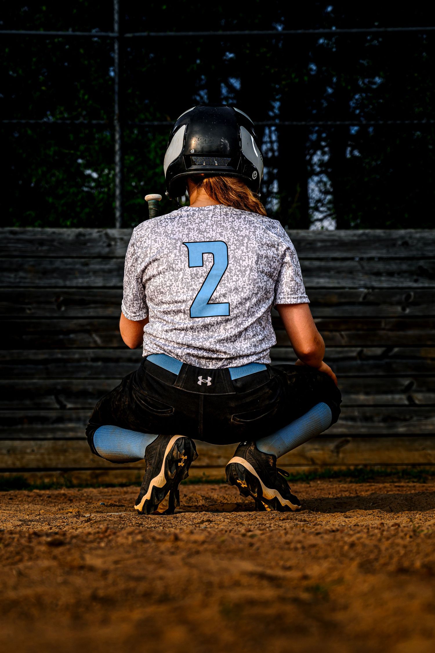 Youth softball player number 2 crouches on dirt field in Under Armour uniform.