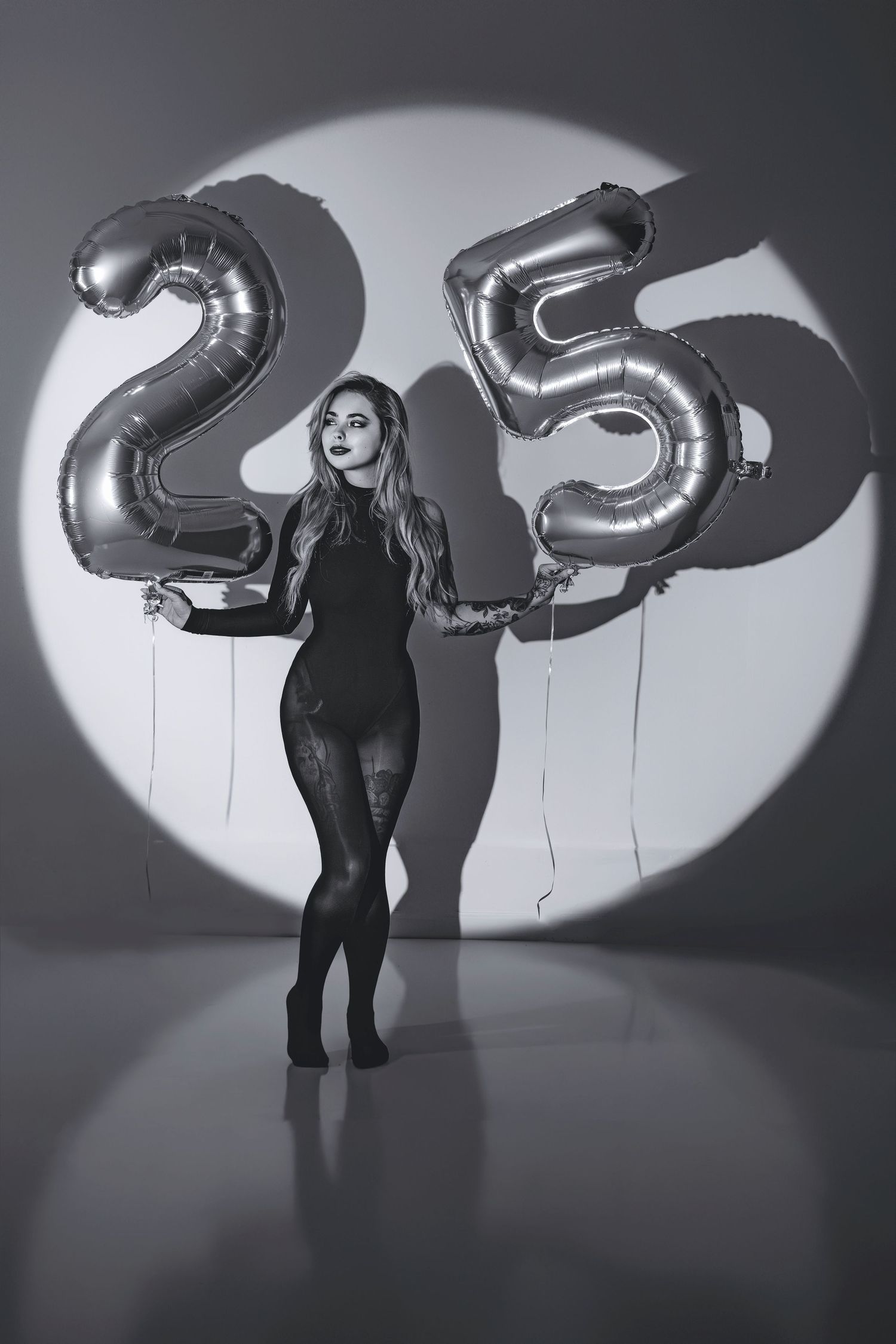 Woman in black outfit holding silver number 25 balloons in dramatic spotlight, black and white birthday photo.