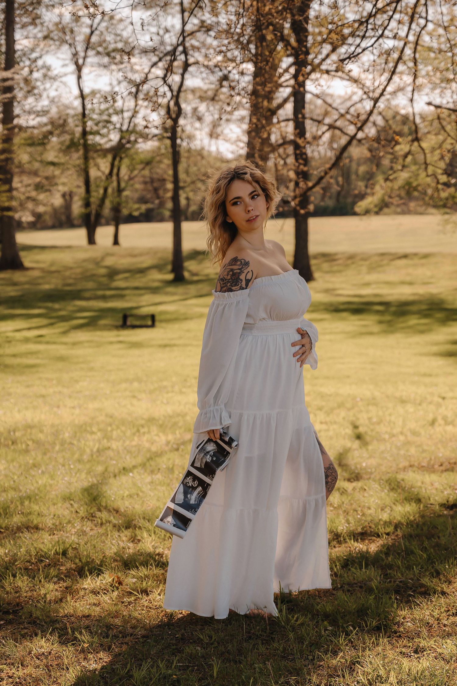 Pregnant woman in white off-shoulder dress holding ultrasound photos in sunny park.