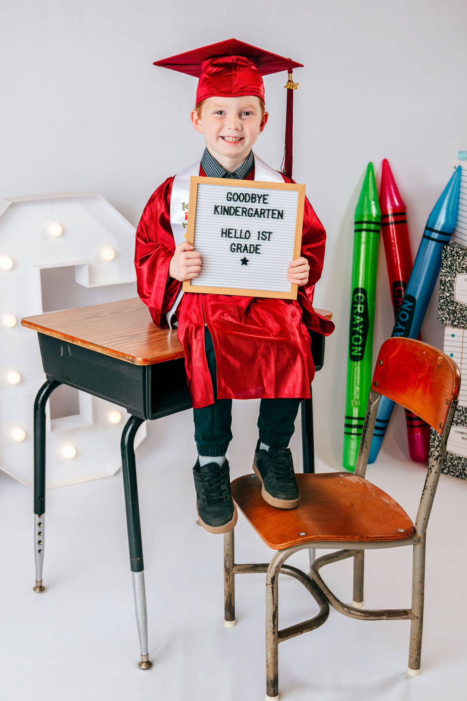 Boy in red graduation cap and gown holds sign reading Goodbye Kindergarten Hello 1st Grade near school desk.