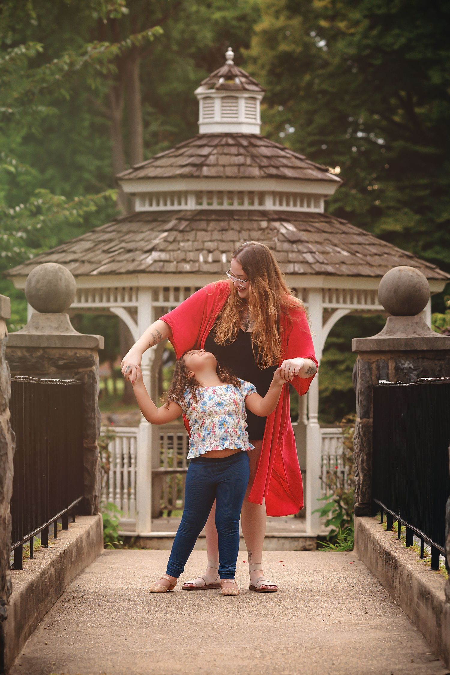 A woman in a red cardigan dances with a young girl on a bridge path near a white gazebo in a park.