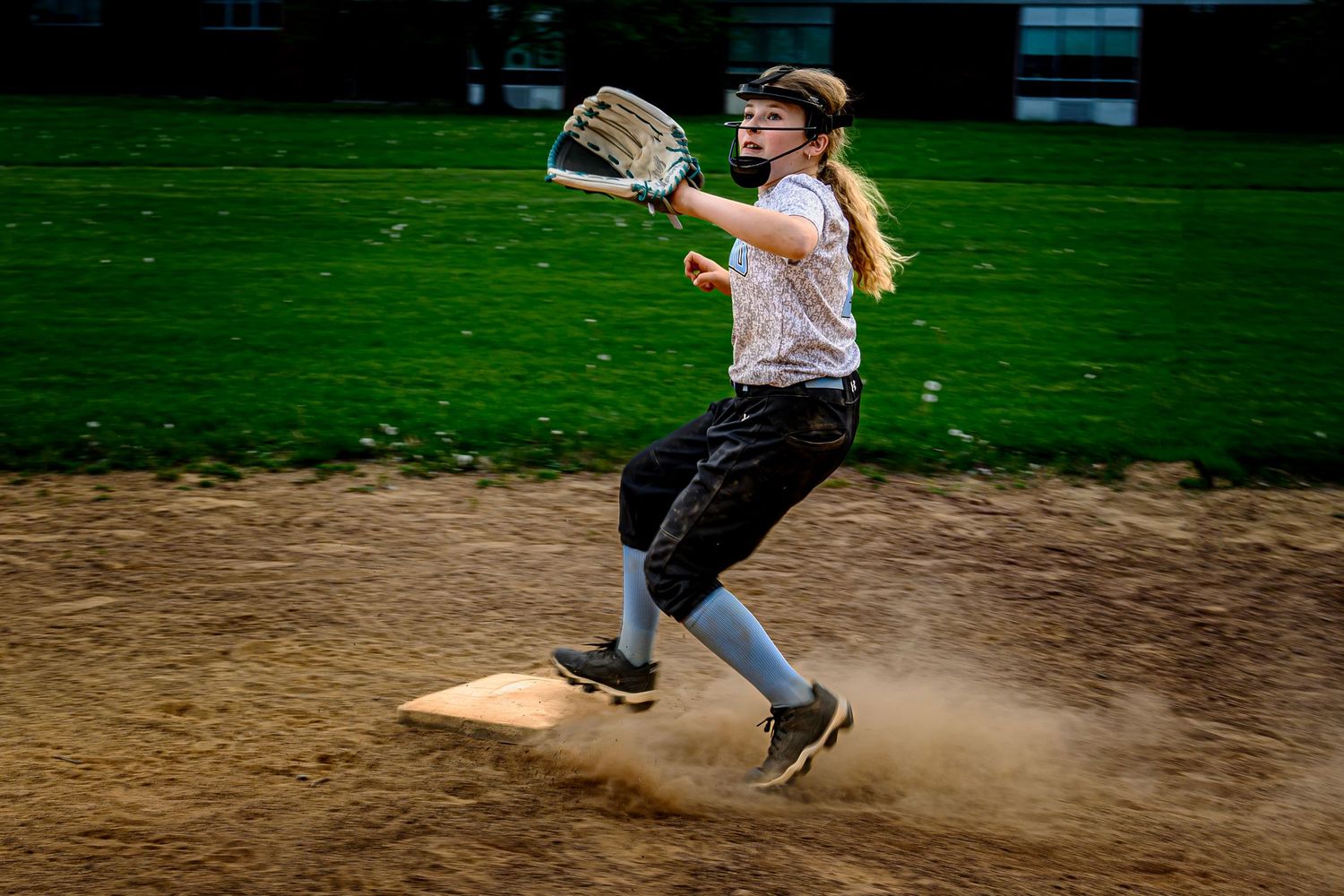 Female softball player catches ball in glove while running past base, kicking up dust on dirt infield.