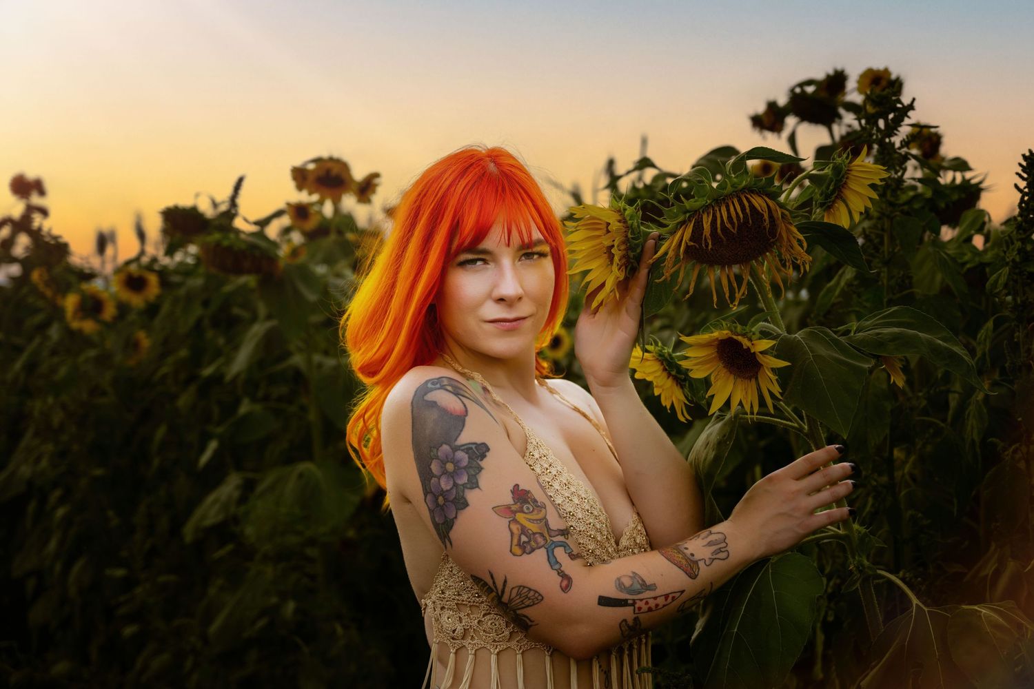 Woman with vibrant orange hair and tattoos poses among sunflowers in a golden sunset field.