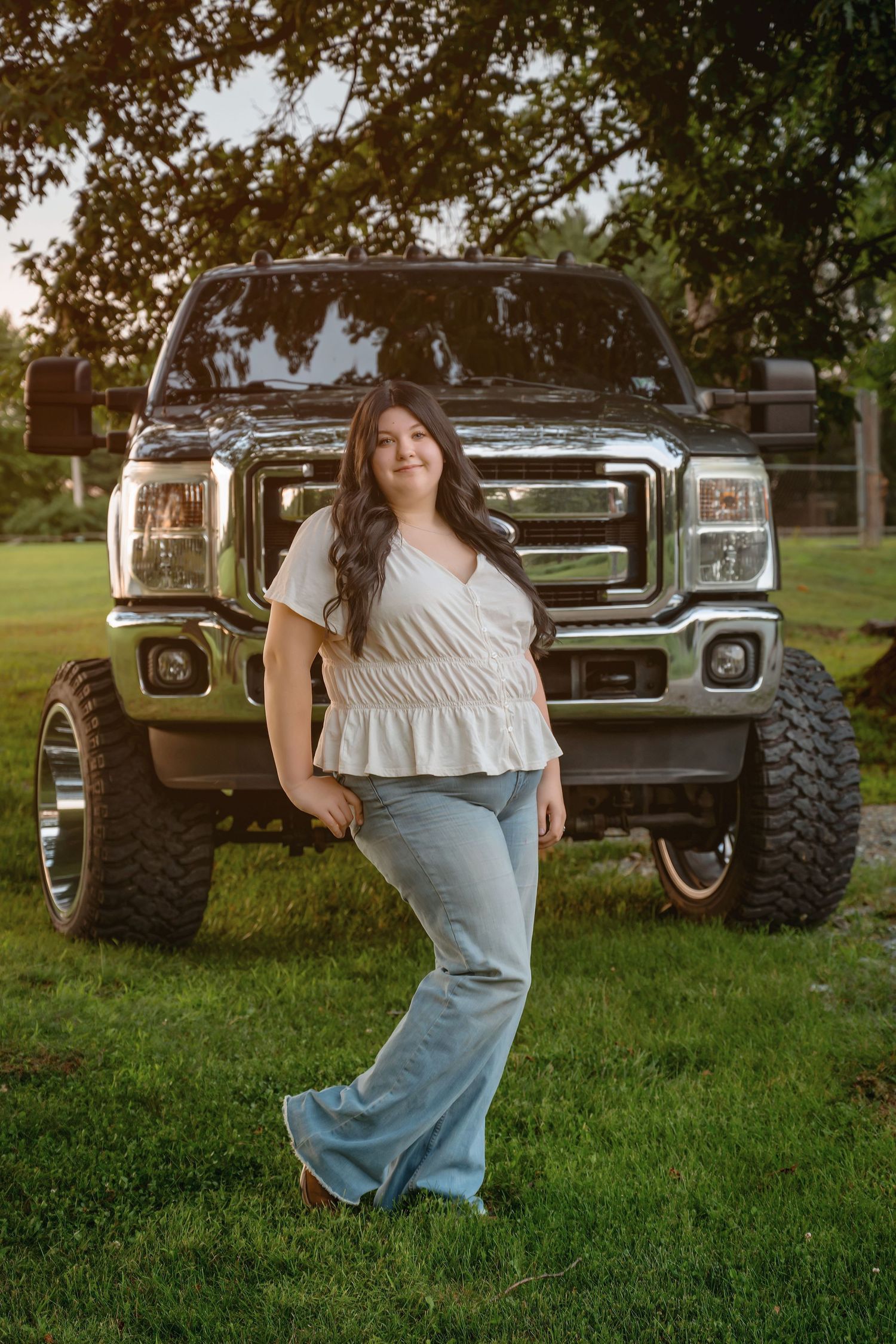 Smiling young woman in white top and jeans posing beside large lifted Ford truck.