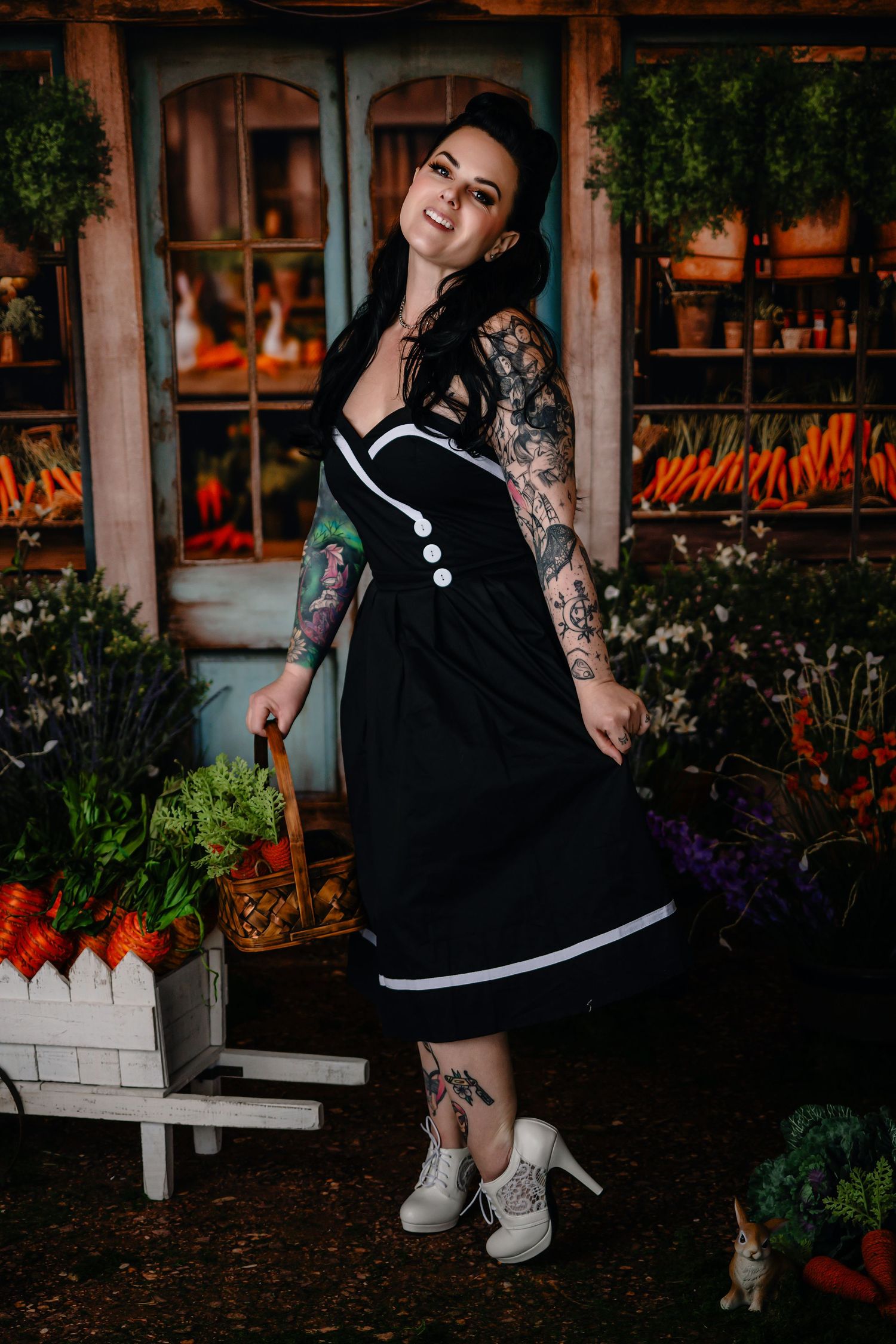 Tattooed woman in black vintage dress with white trim posing in a colorful garden shop setting.