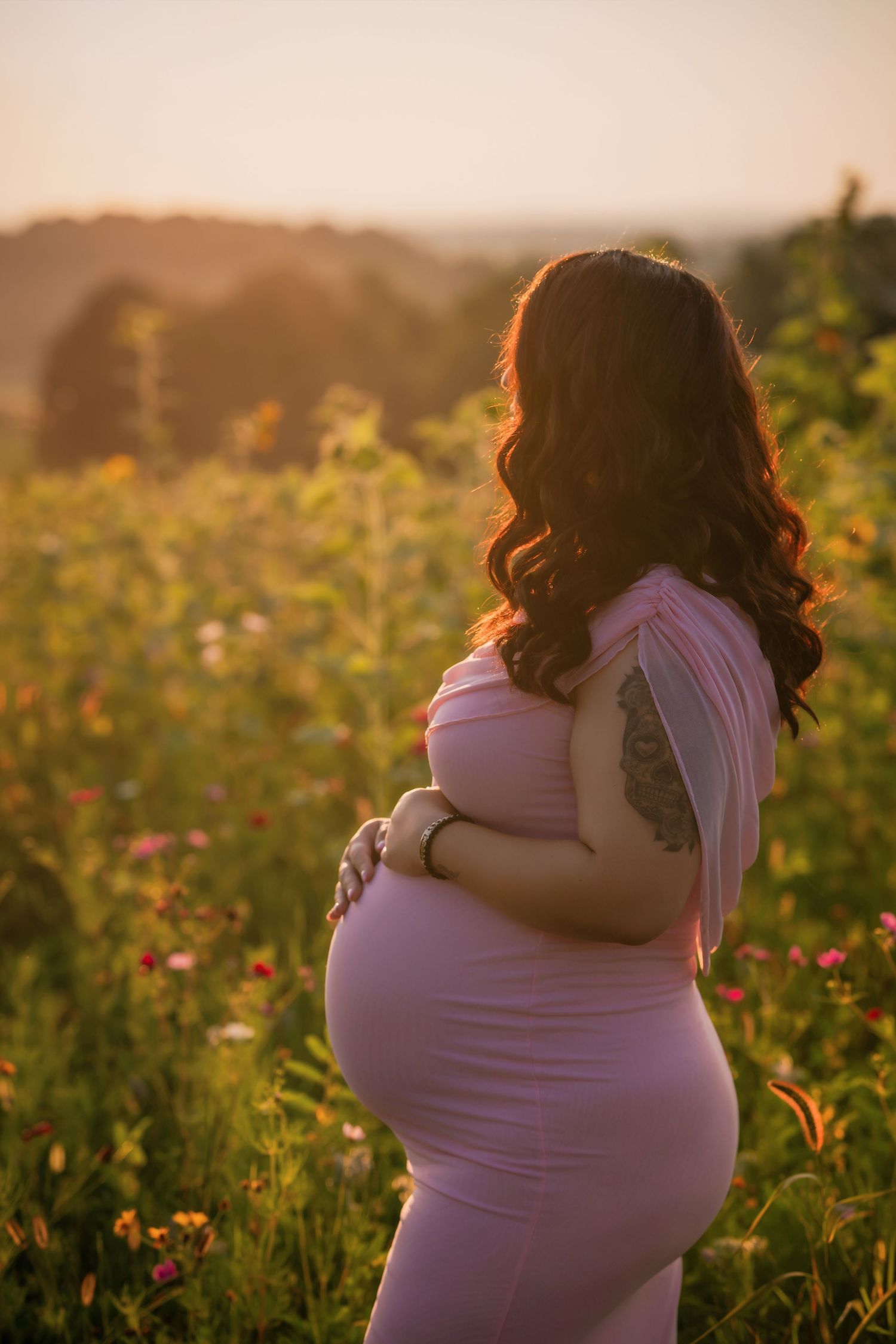 Pregnant woman in pink dress holding baby bump in sunlit flower field at golden hour.