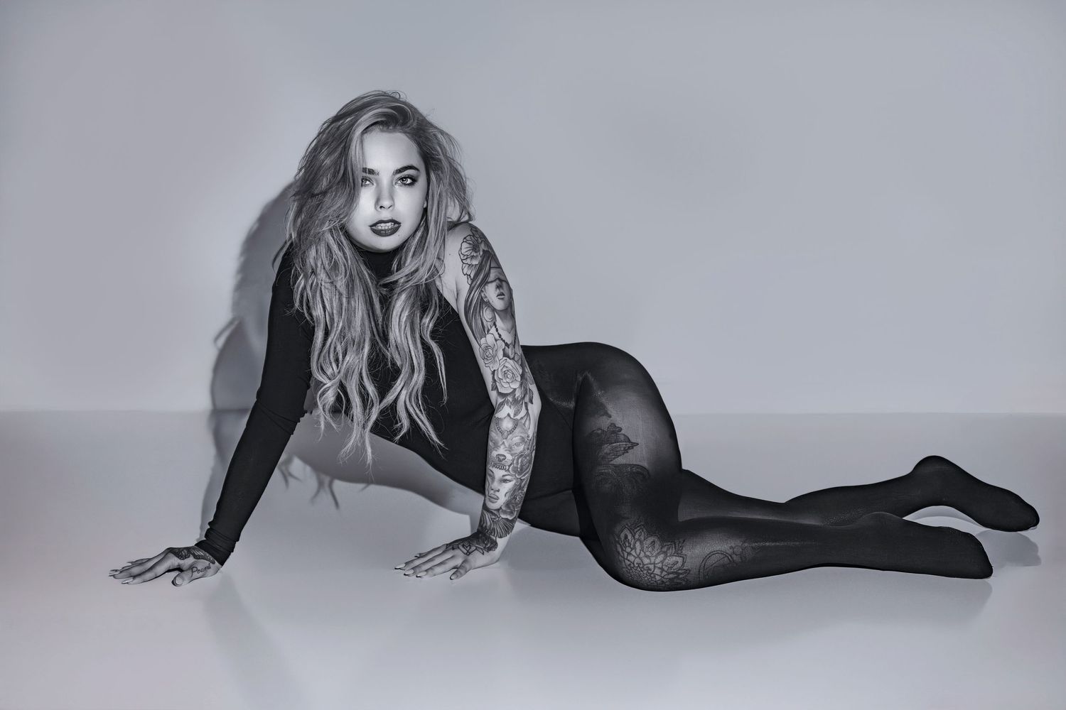 Black and white photo of a tattooed woman in a sheer bodysuit posing elegantly on the floor.