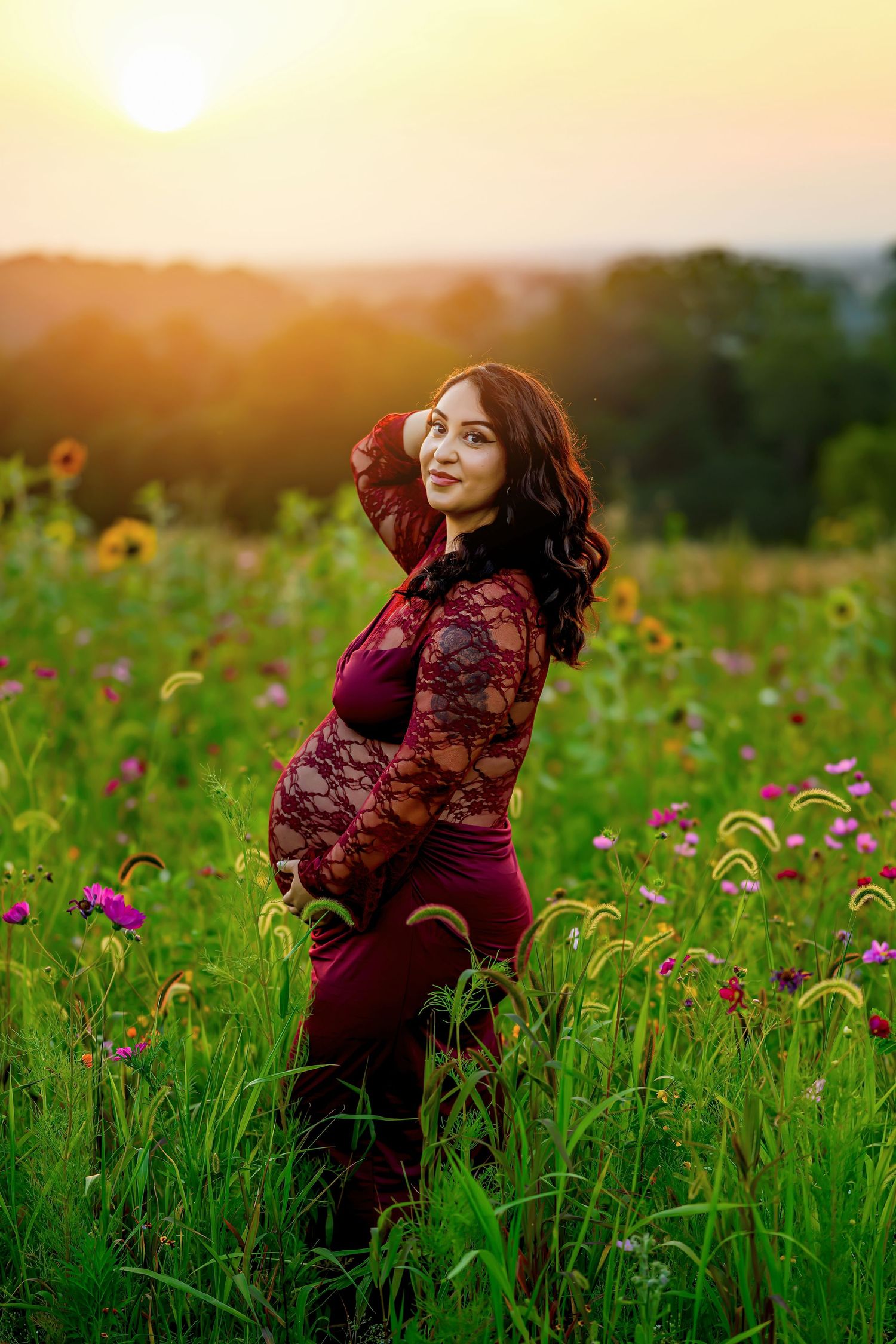 Smiling pregnant woman in burgundy lace dress posing in colorful wildflower field at sunset.