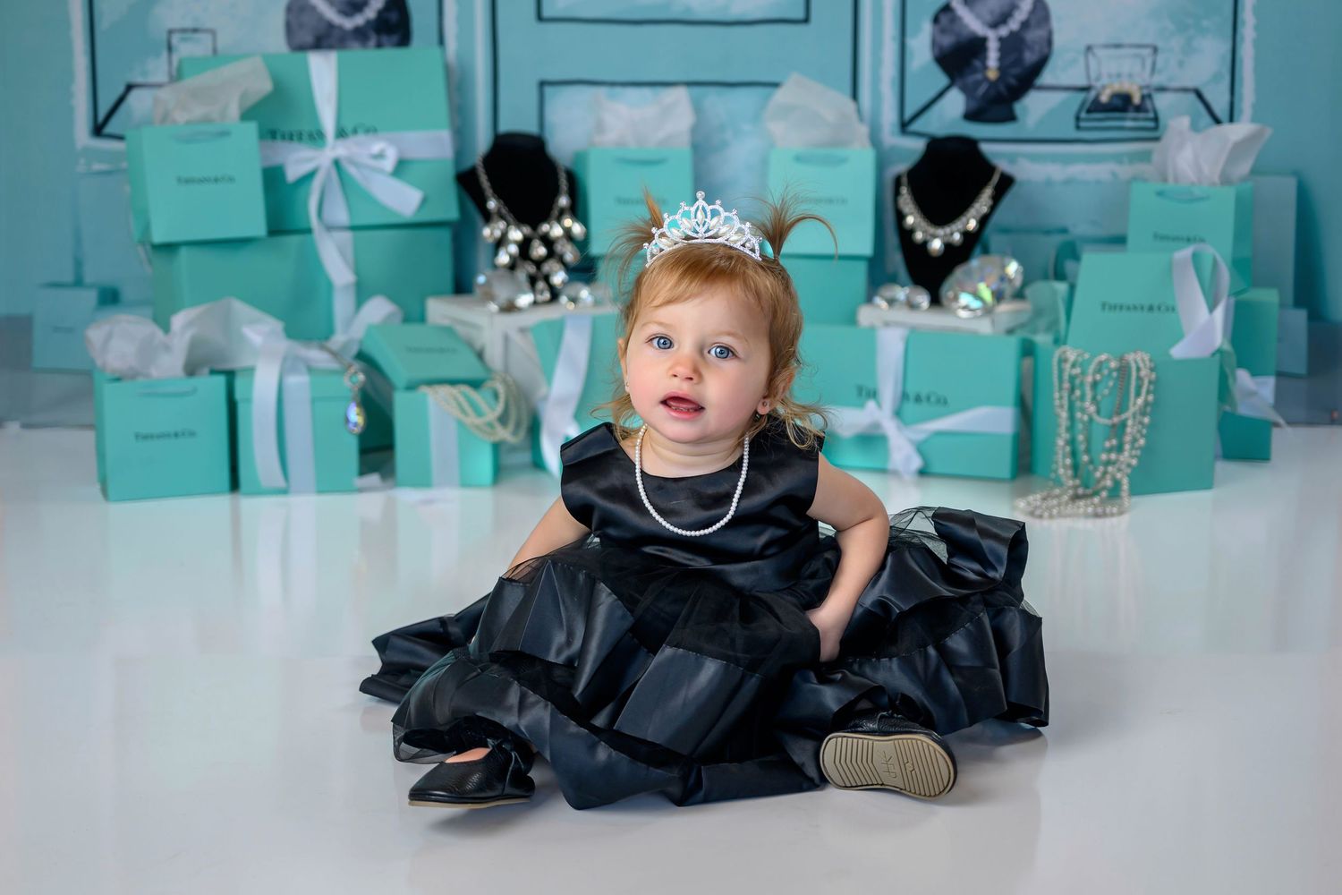 Elegant baby girl in black dress with pearl necklace and tiara surrounded by teal gift boxes.
