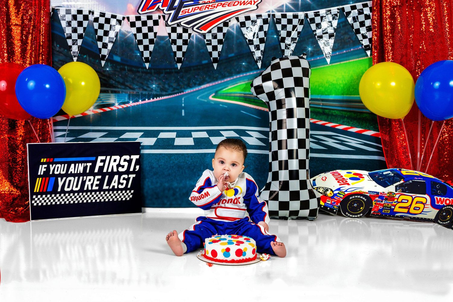 Baby in racing suit smashes first birthday cake beside NASCAR-themed decorations.