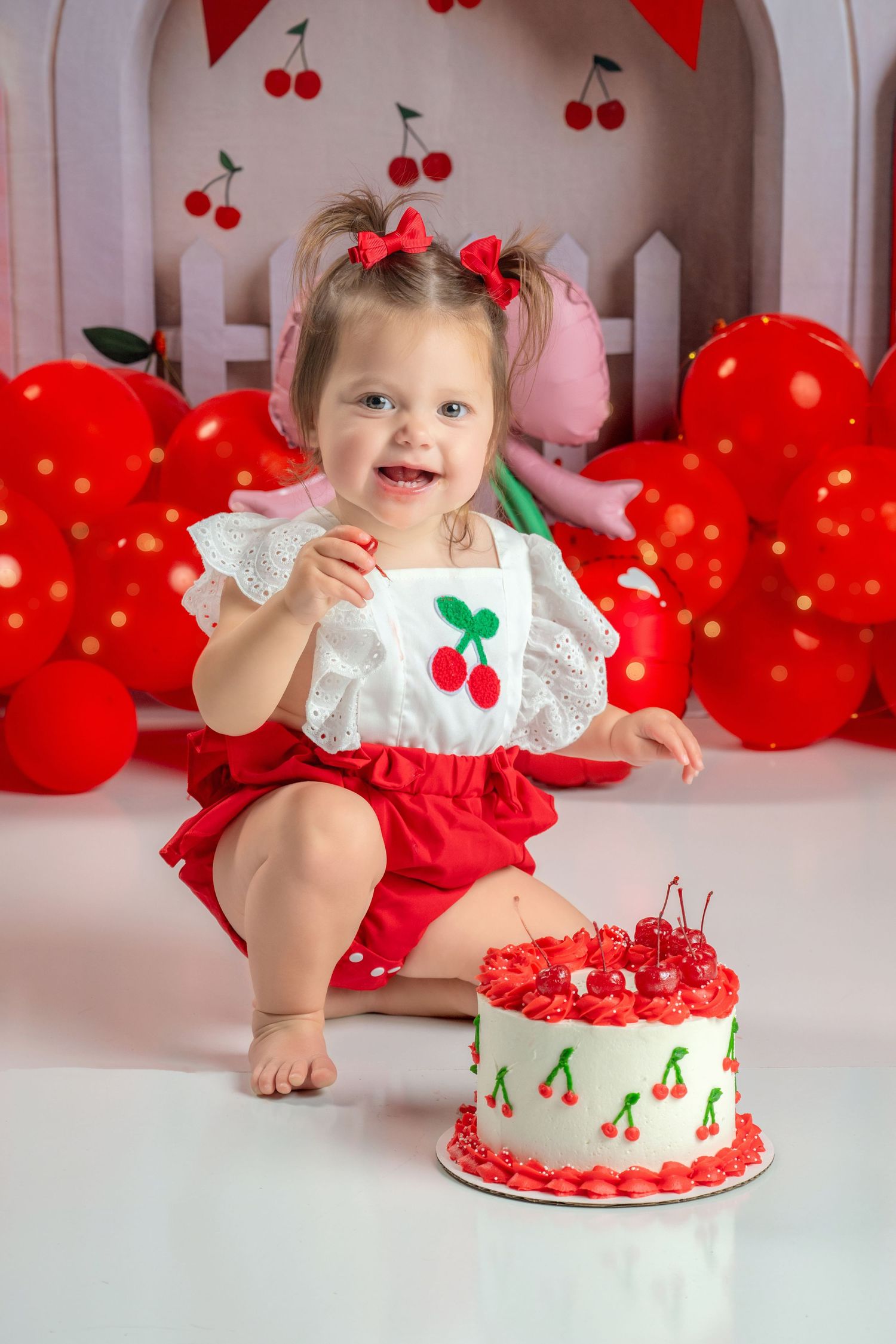 Happy toddler girl in cherry outfit celebrating first birthday with decorated cherry smash cake.