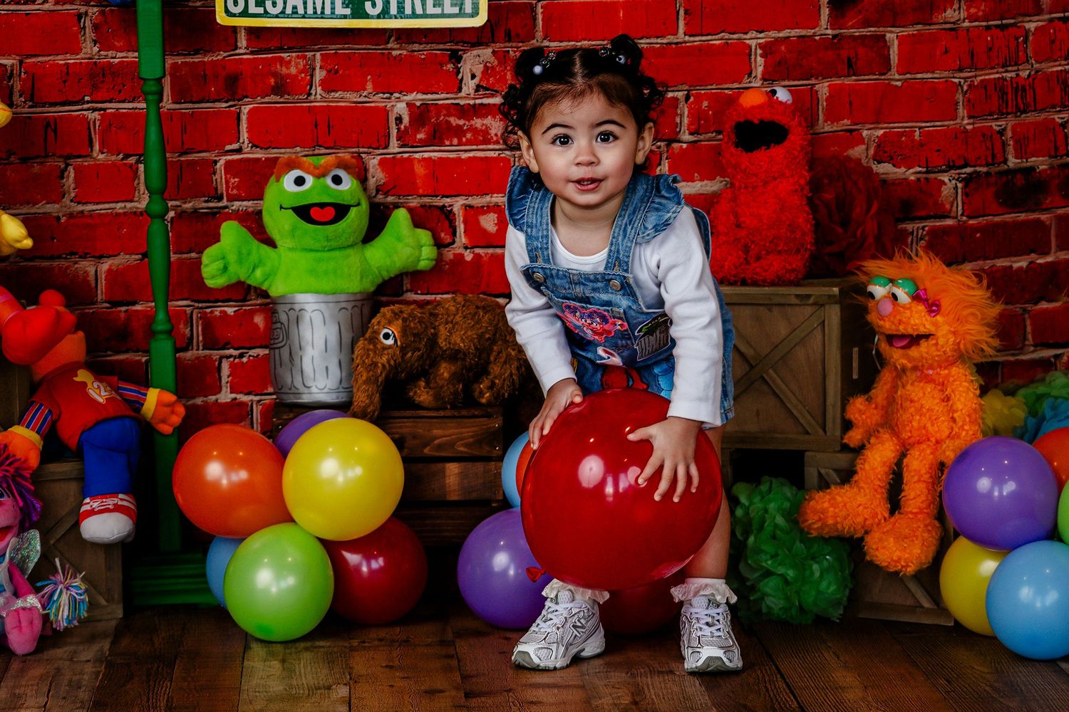 Toddler girl in denim overalls plays with red balloon amid Sesame Street character toys.