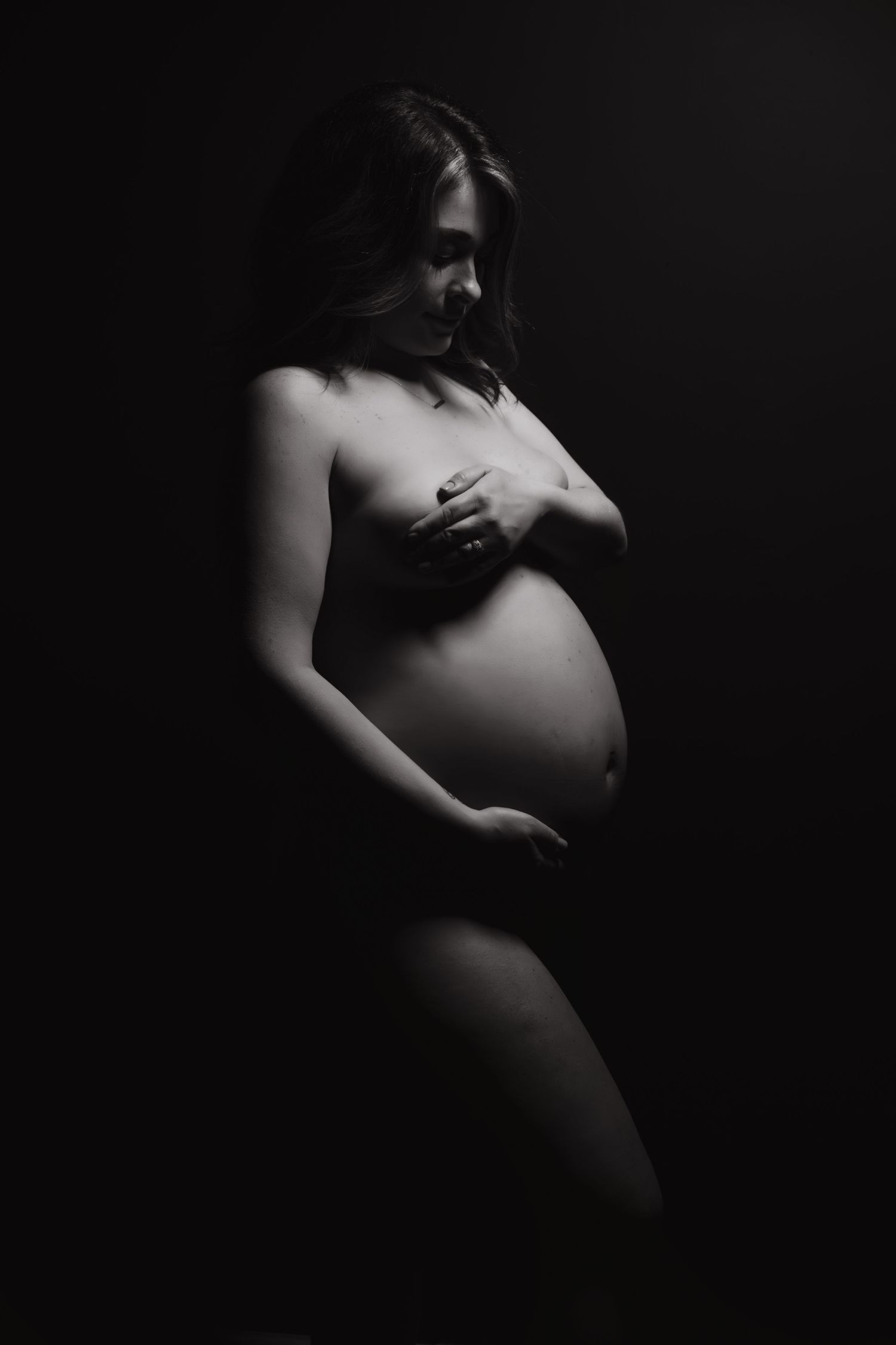 Black and white artistic maternity photo of pregnant woman in profile pose against dark background.