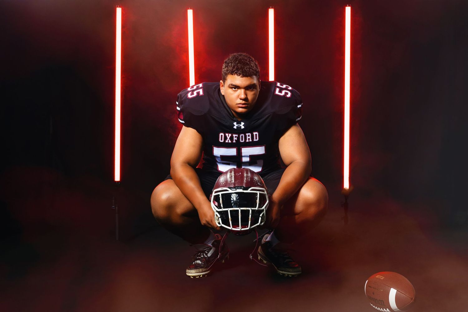 Oxford #55 football player crouching with helmet in red neon lit studio.