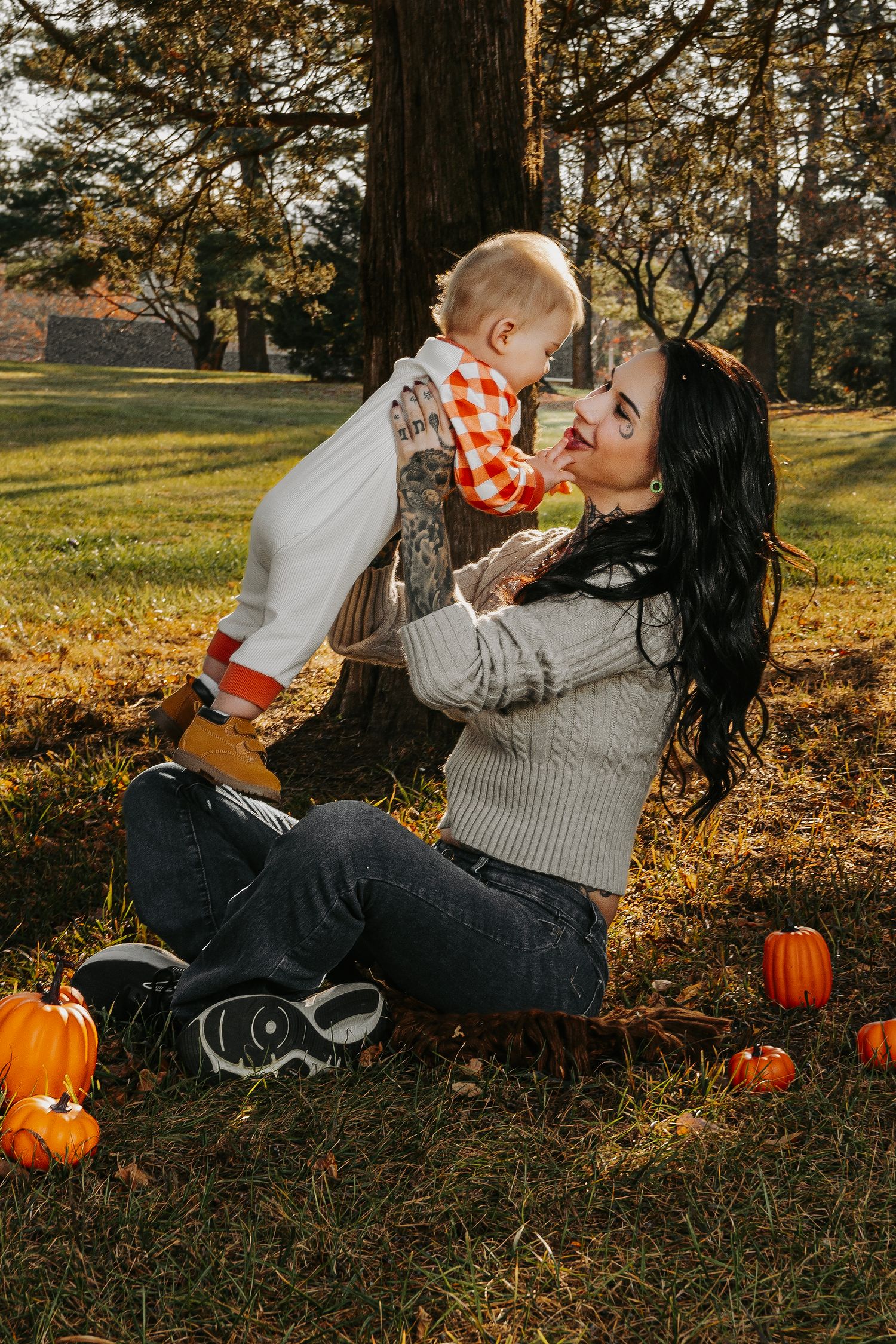 A mother sits on autumn grass holding her baby up, surrounded by small pumpkins near a tree in golden fall light.