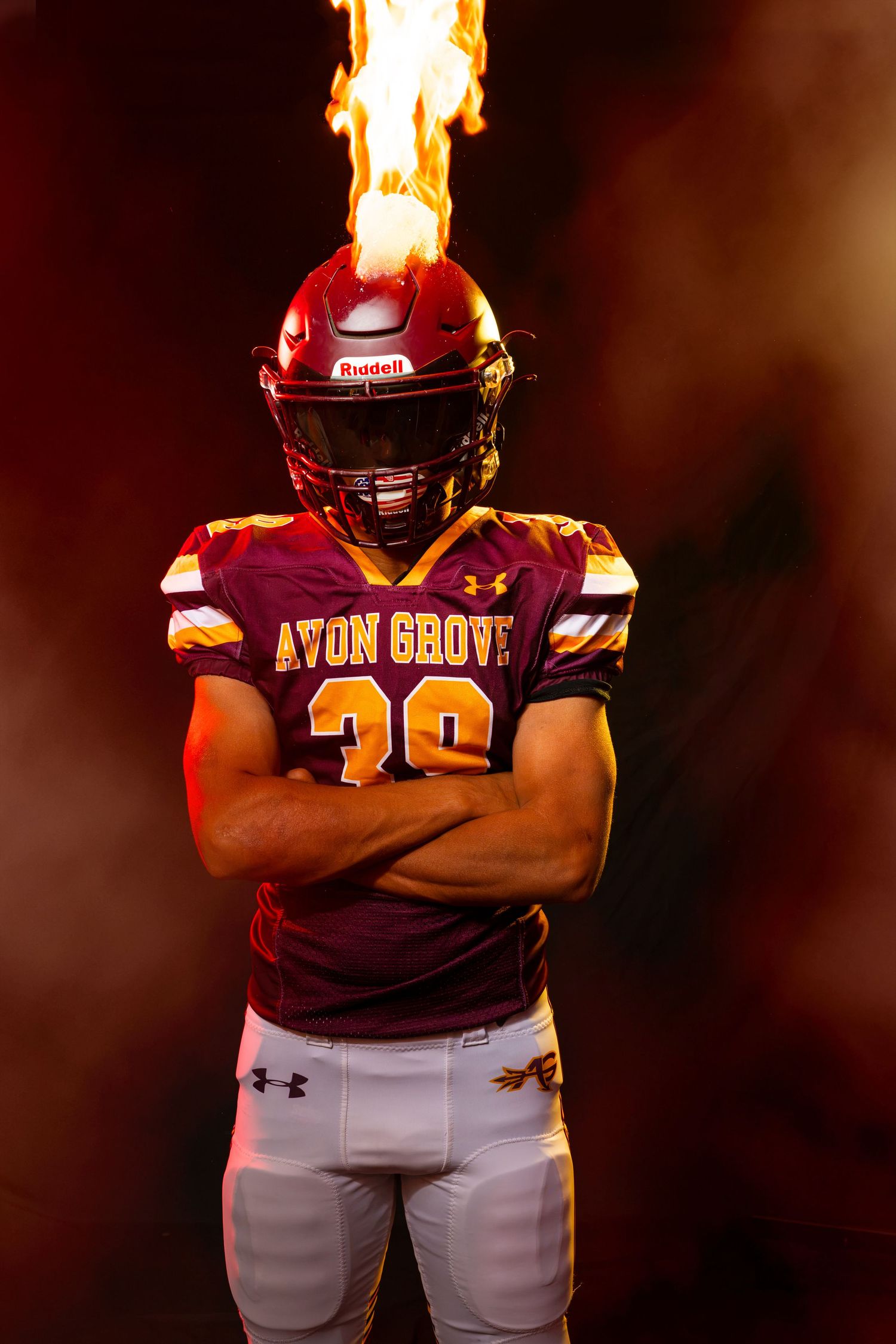 Avon Grove football player #39 in maroon uniform stands with arms crossed as flames erupt from helmet.