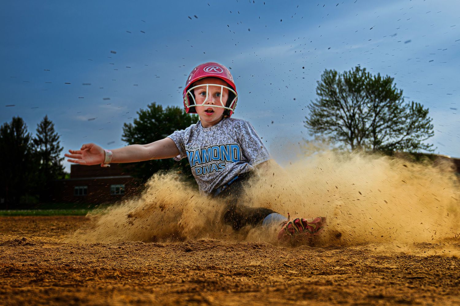 Young softball player in pink helmet slides into base sending dirt flying in dramatic action shot.