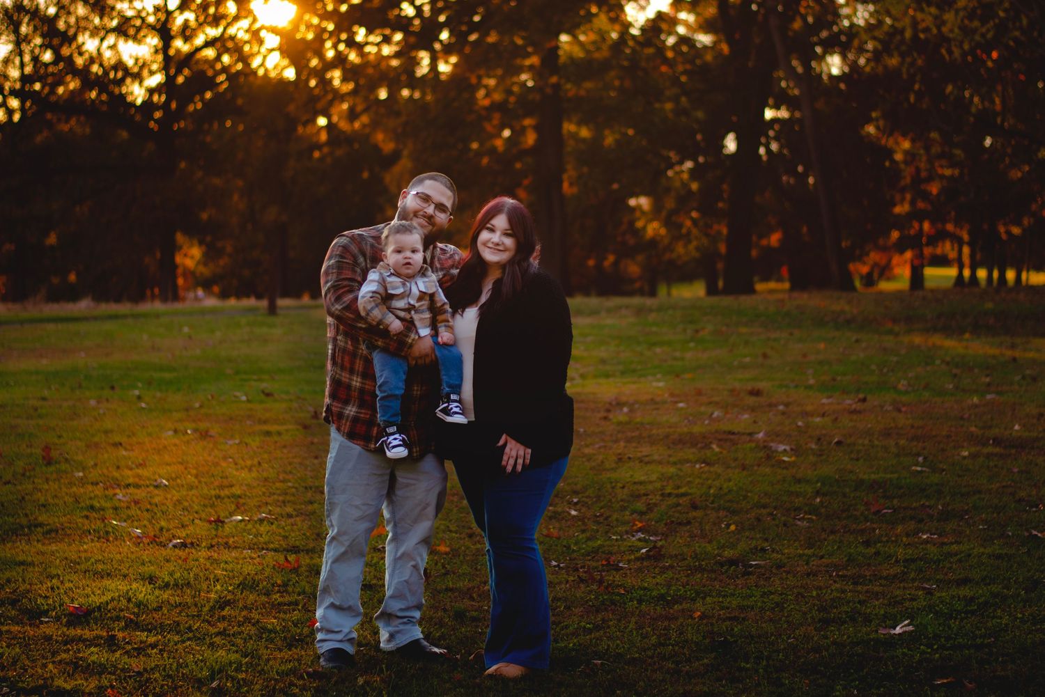 Family of three posing at golden hour in a park, parents holding baby amid autumn foliage.