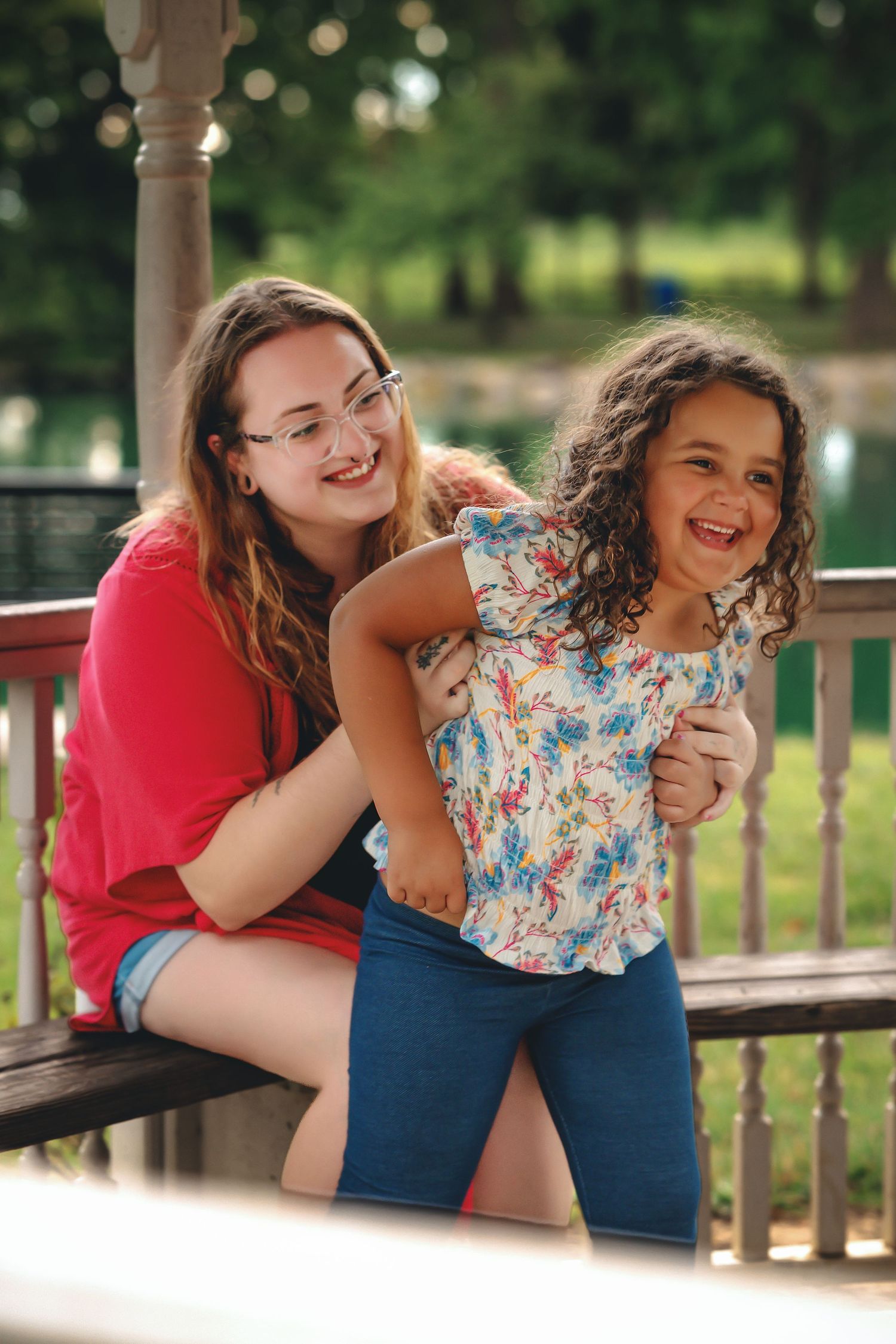 Two girls laughing and playing together on a wooden bench in a sunny park setting.