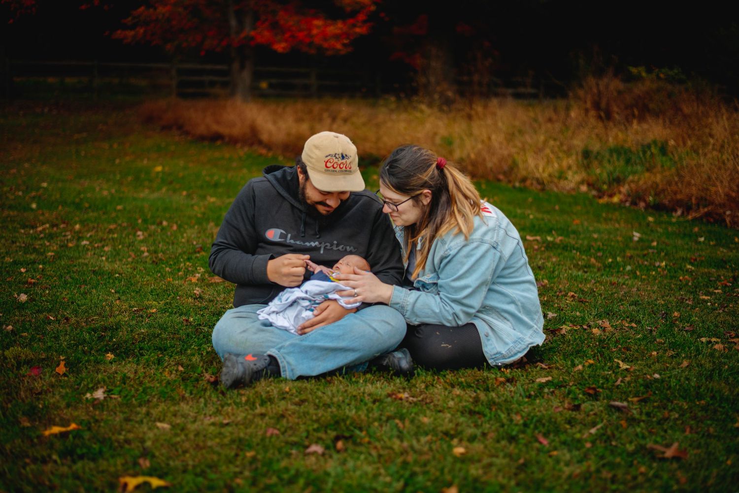 A couple sits on autumn grass, lovingly looking at their newborn baby wrapped in a light blue blanket.