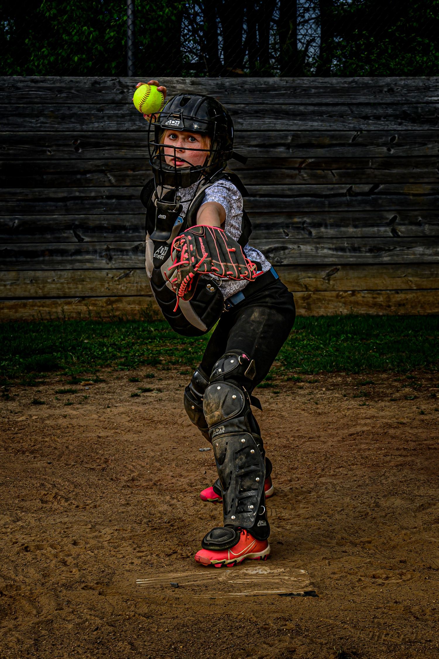 Young softball catcher in full gear reaches for incoming yellow ball at home plate.