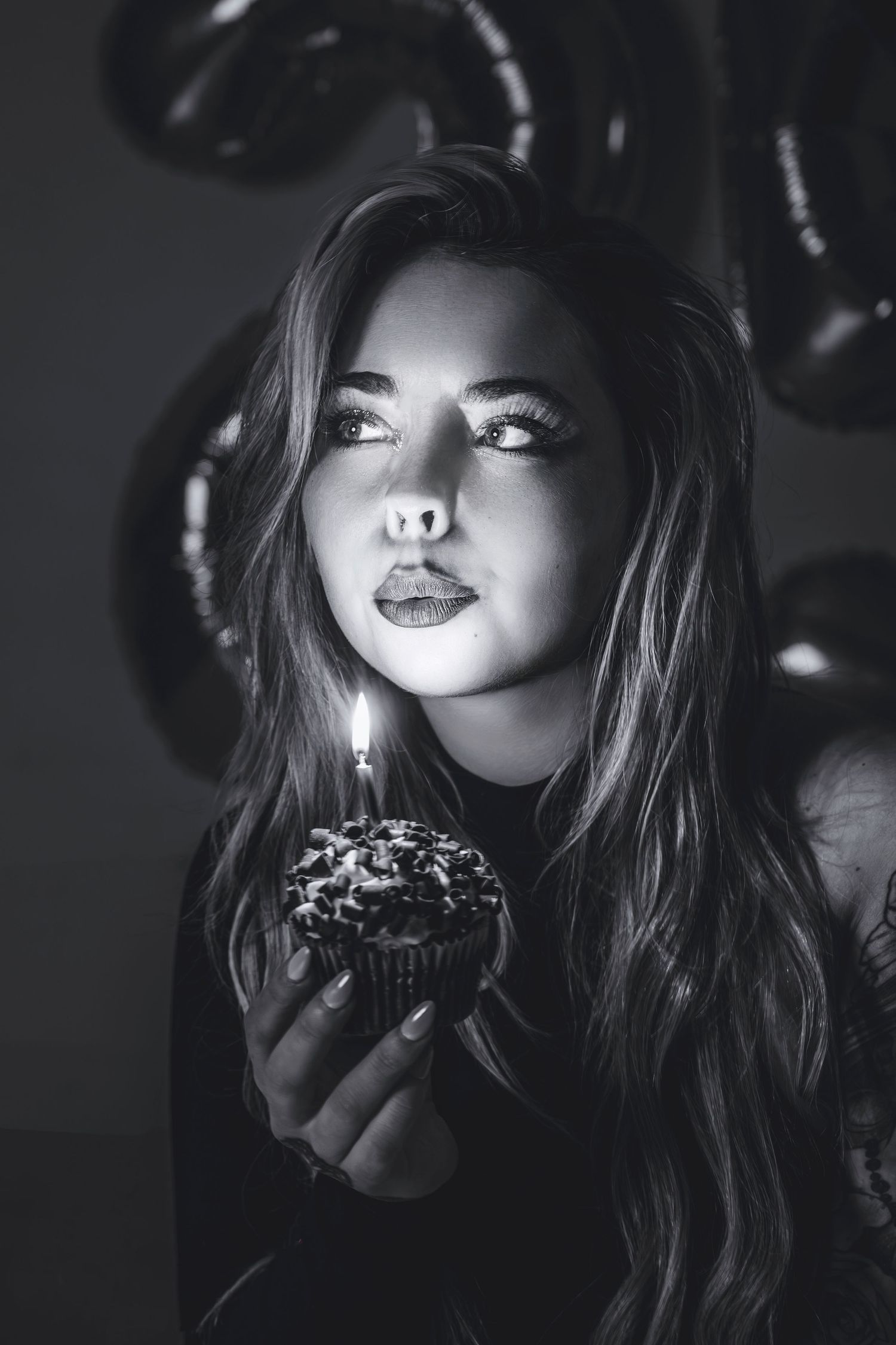 Black and white portrait of young woman blowing out candle on birthday cupcake with balloons.
