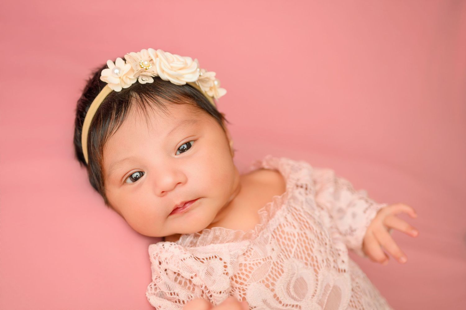 Newborn baby girl in white lace dress and floral headband lying on pink backdrop.