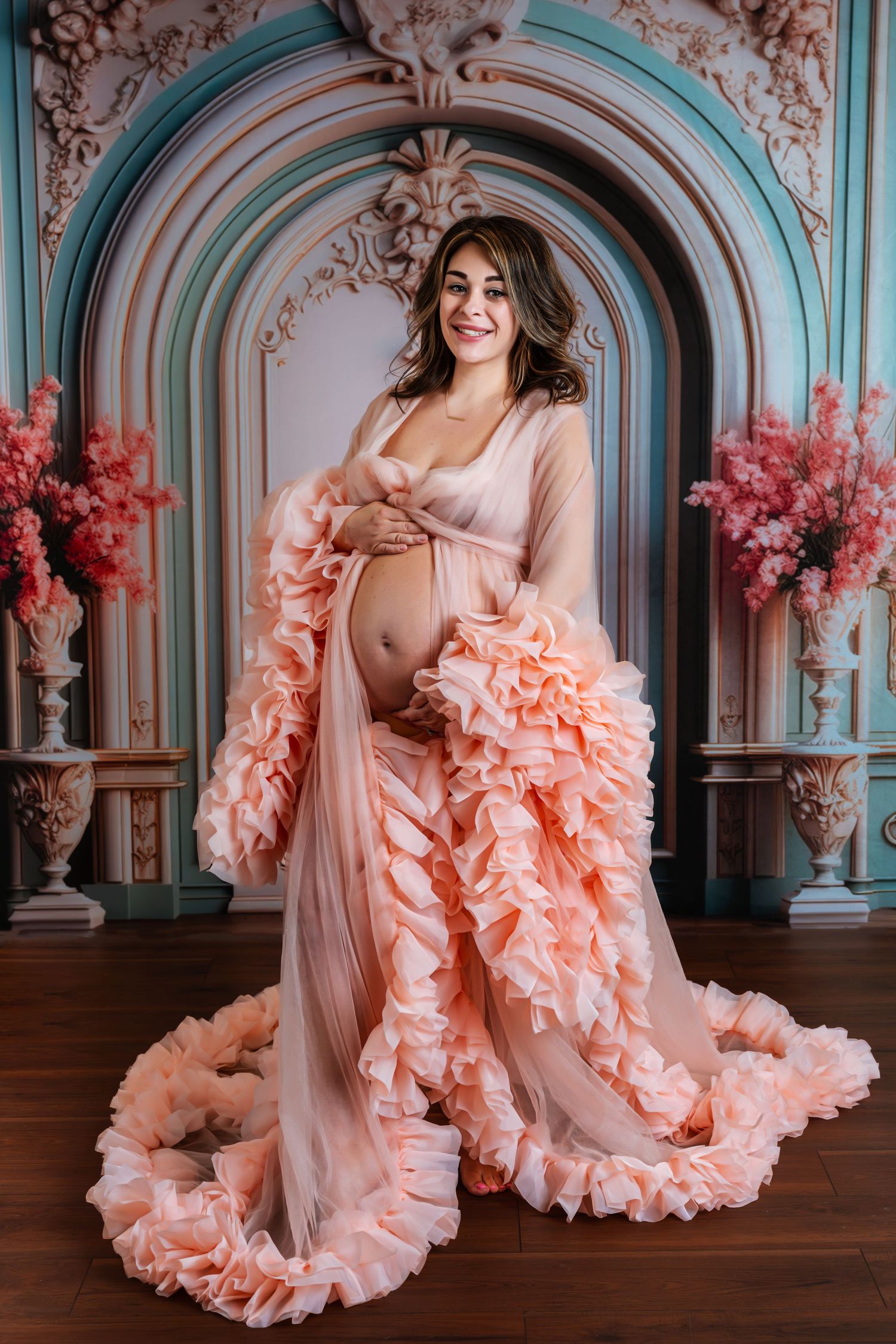Smiling pregnant woman in elegant ruffled peach gown posing in ornate decorative studio setting.