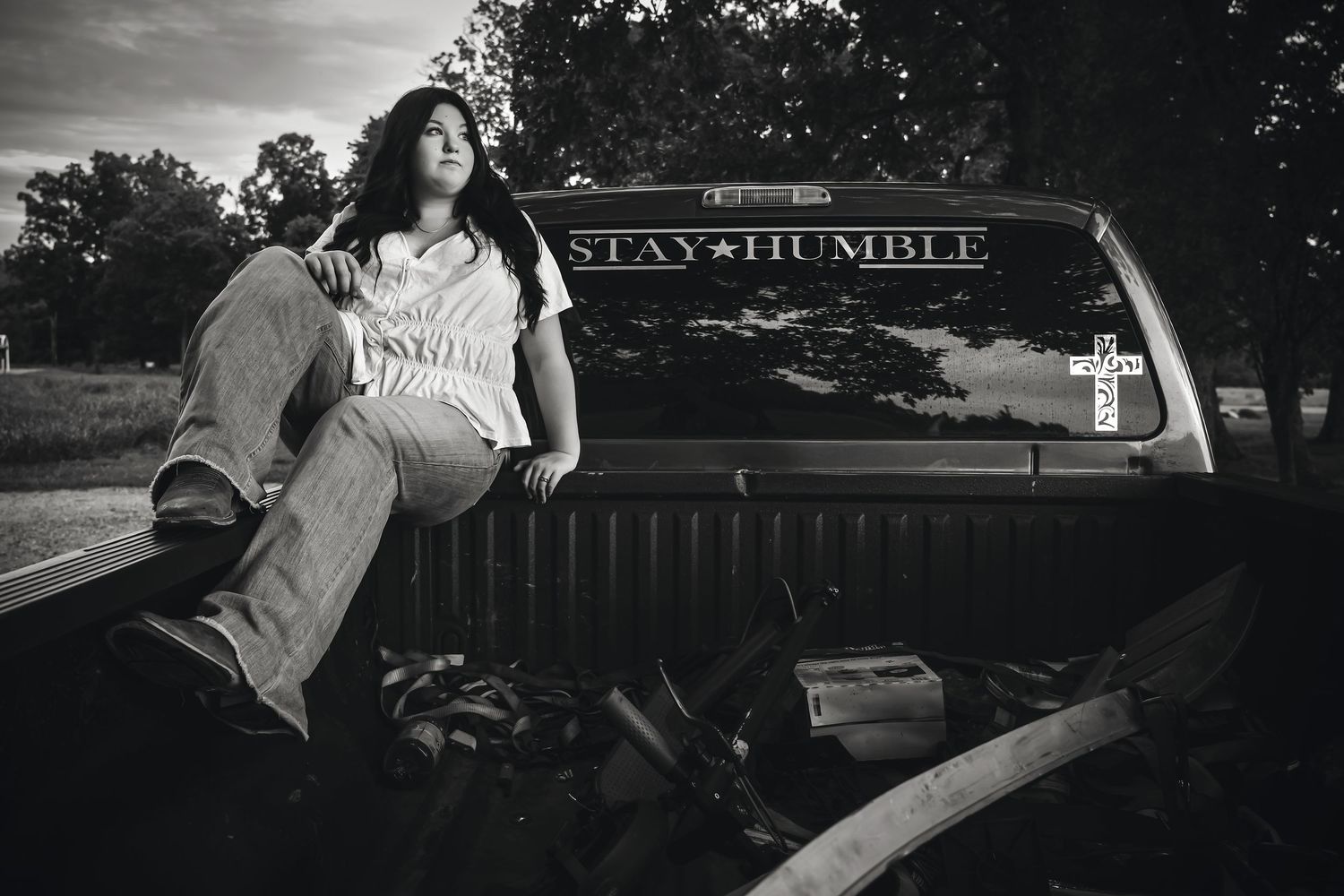 Black and white photo of a young woman sitting on a pickup truck tailgate with Stay Humble decal, outdoors.