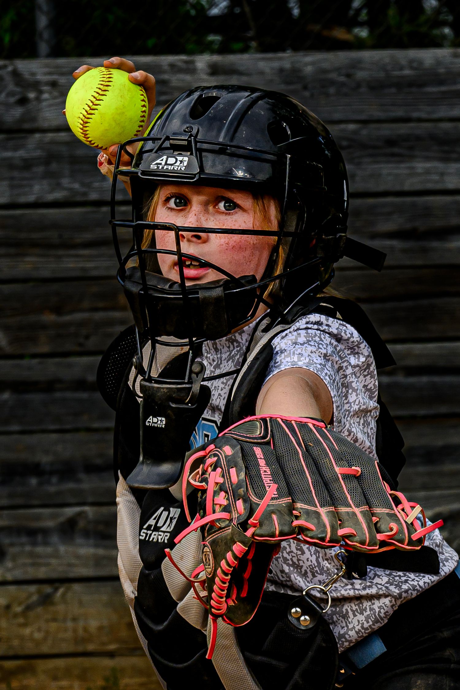 Young softball catcher in black gear holds yellow softball with glove outstretched.
