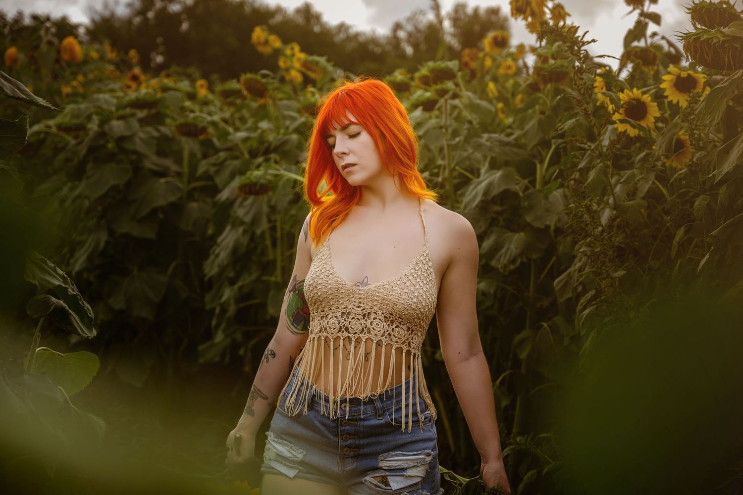 A woman with vibrant orange hair wearing a crochet crop top and denim shorts stands among tall sunflowers.