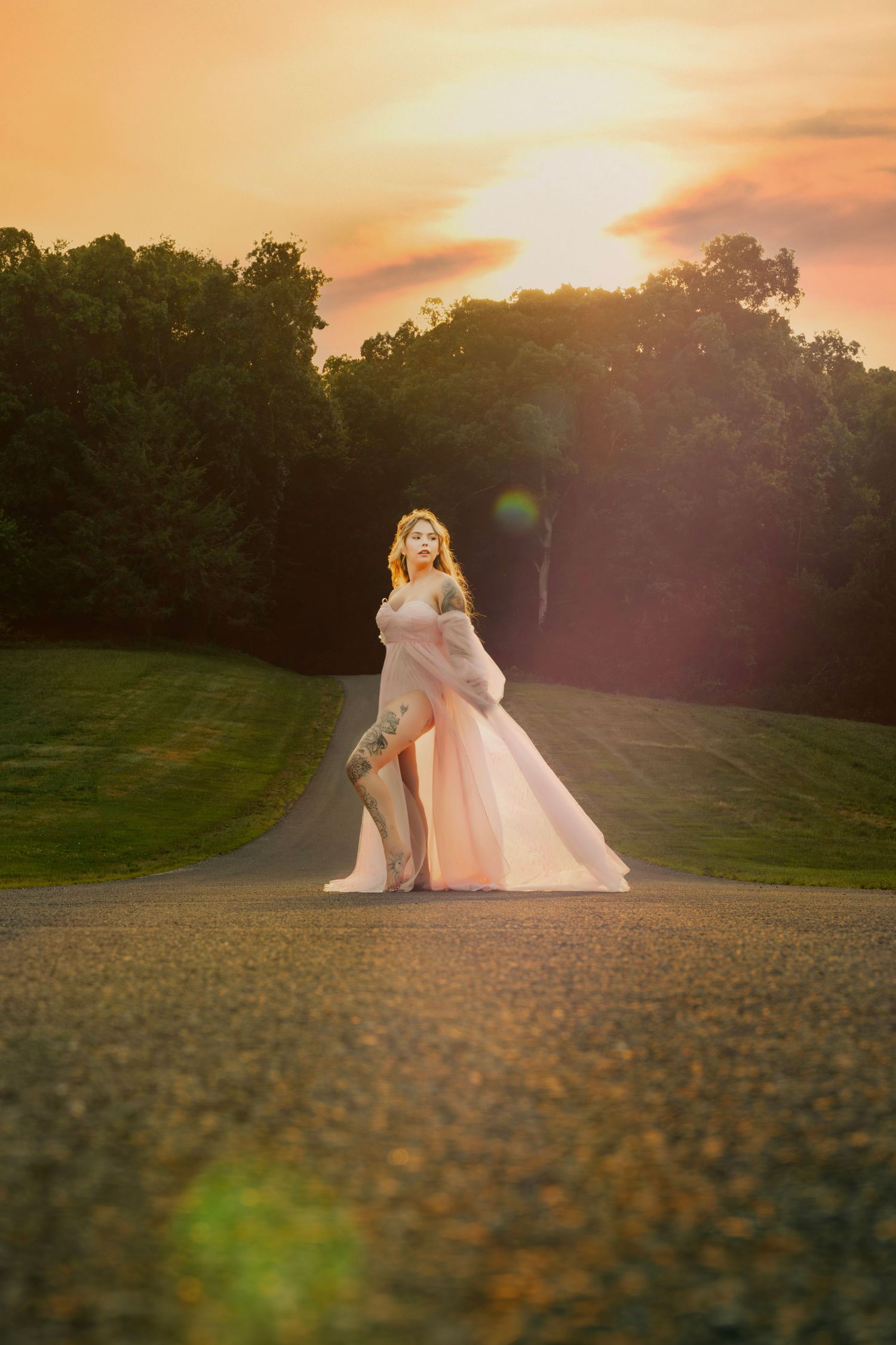 Blonde woman in flowing pink gown standing on a path surrounded by lush greenery at sunset.