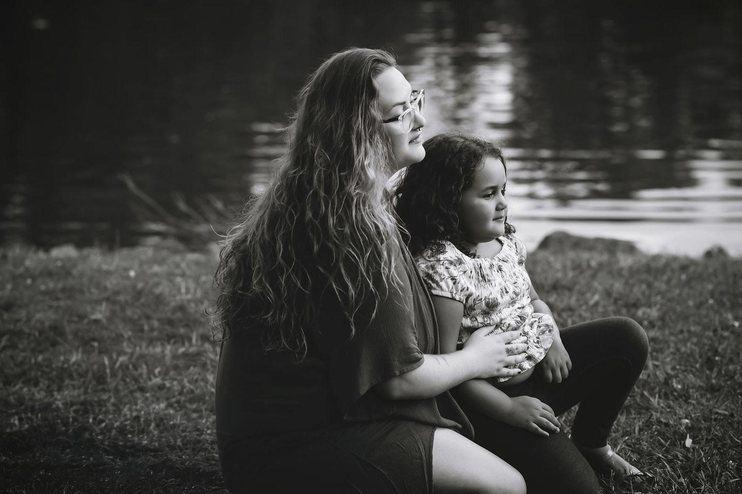 Black and white photo of a woman with long hair holding a young girl by a lake on grass.
