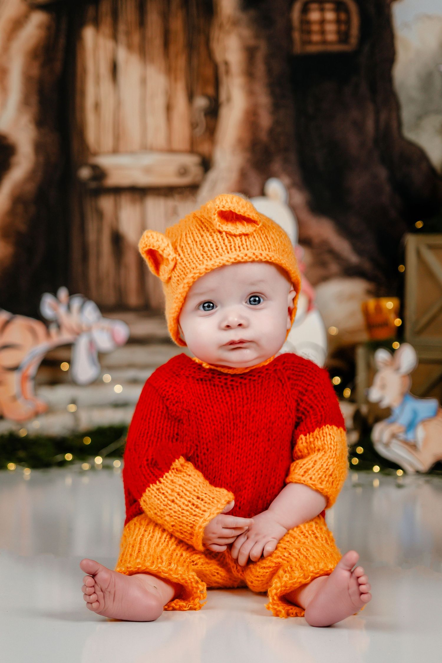 Baby in knitted Winnie the Pooh costume with orange hat and red sweater sits before festive woodland backdrop.