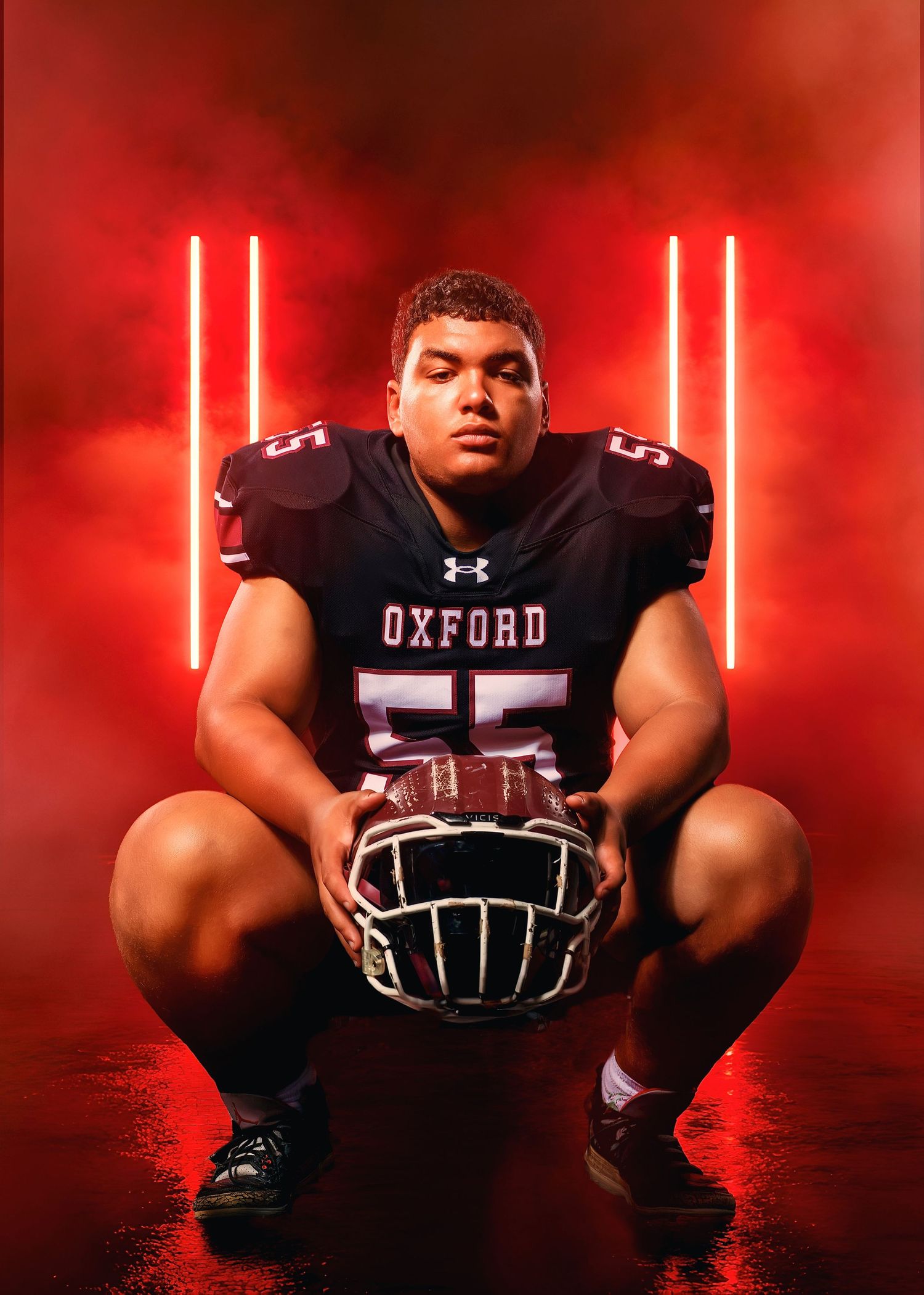 Oxford #55 football player crouches holding helmet in dramatic red smoke studio portrait.