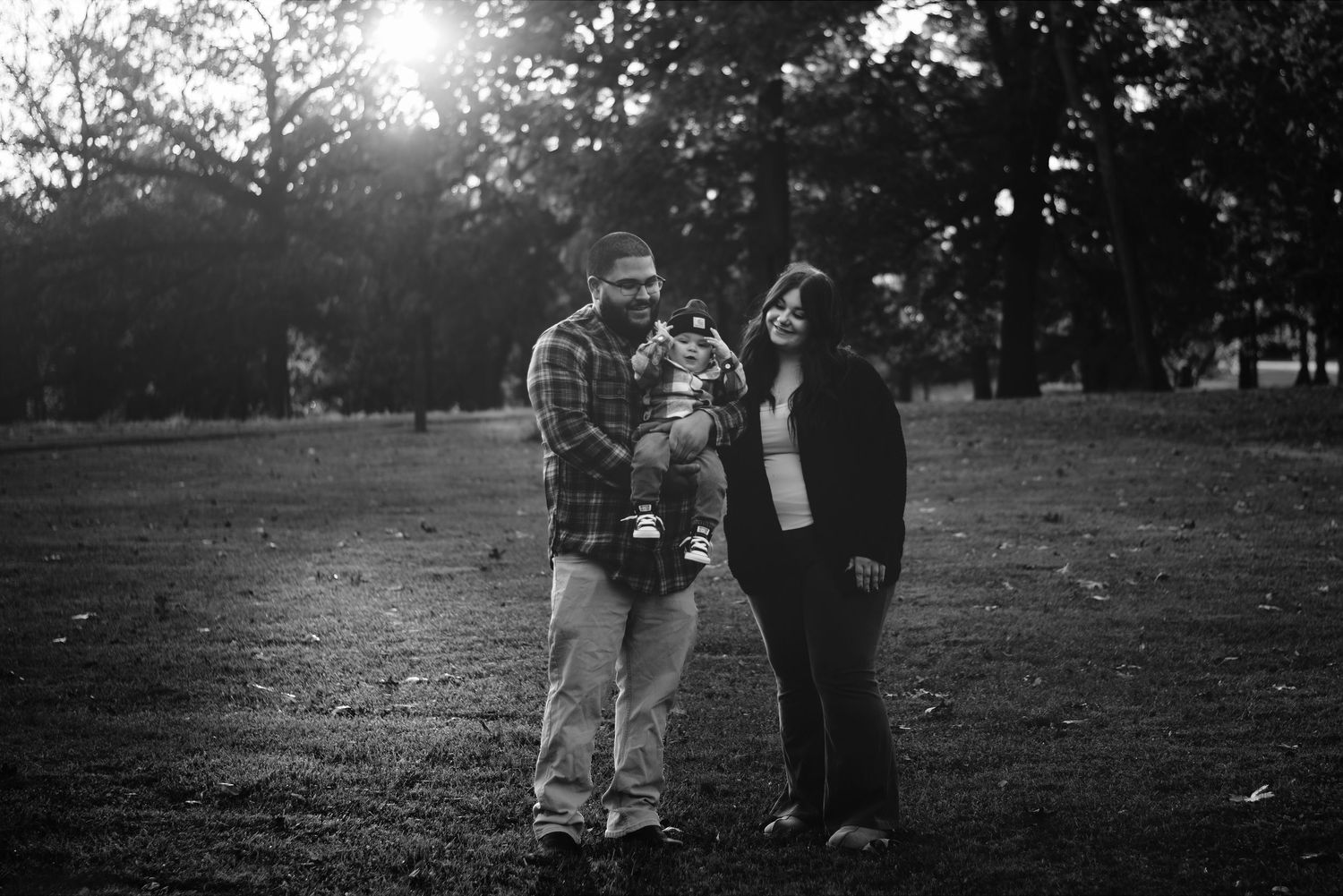 Black and white photo of a couple holding their baby in a sunlit park surrounded by trees.