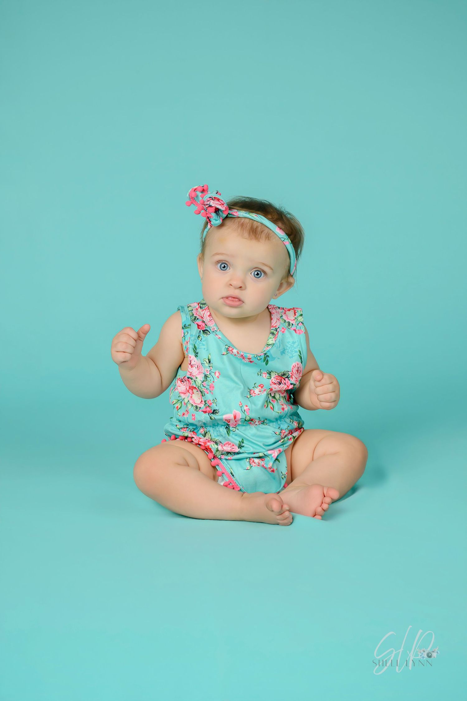Baby girl in teal floral romper and matching headband sits on teal backdrop for professional portrait photo.