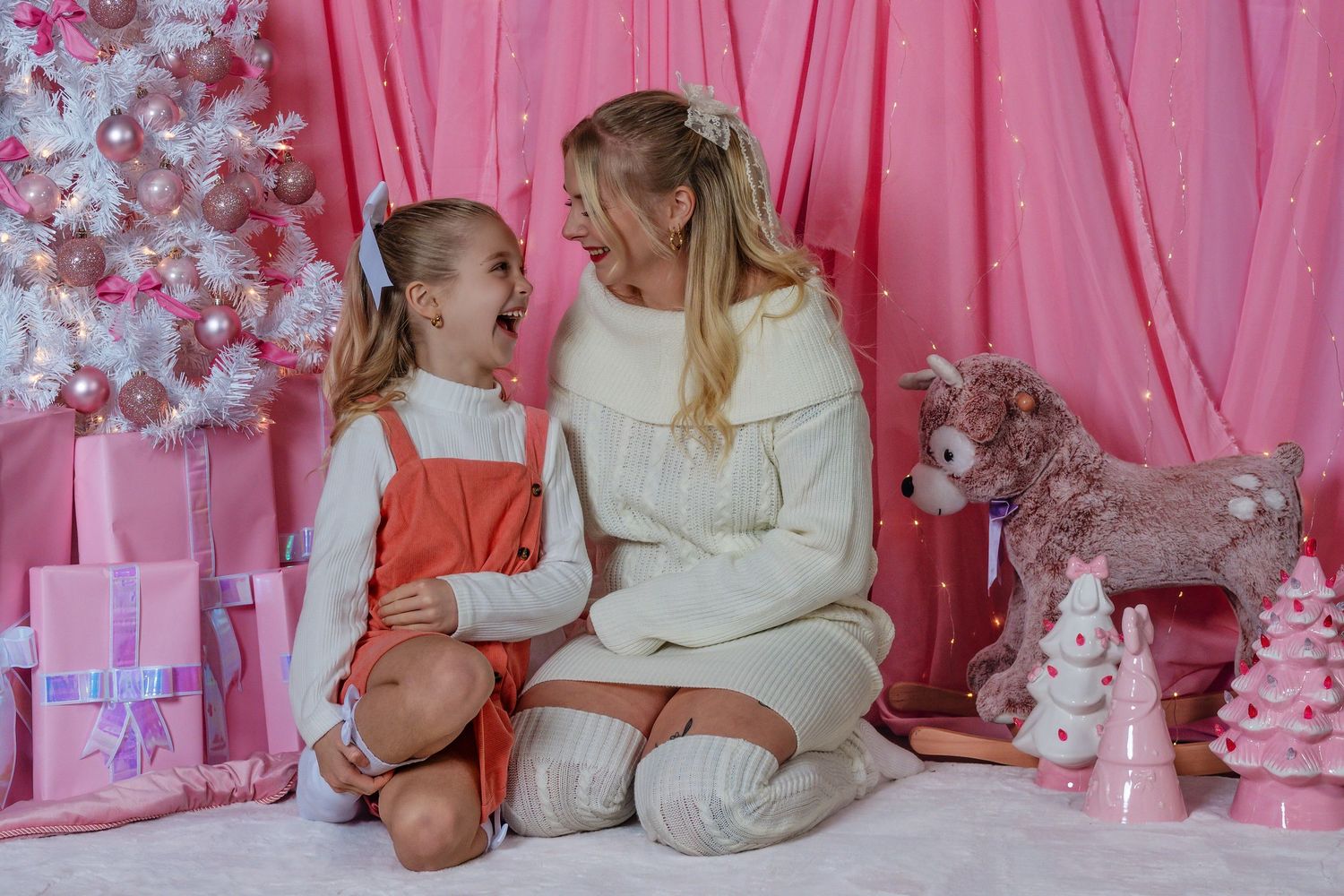 Mom and daughter share a joyful moment in a pink Christmas setting with gifts and a white tree.
