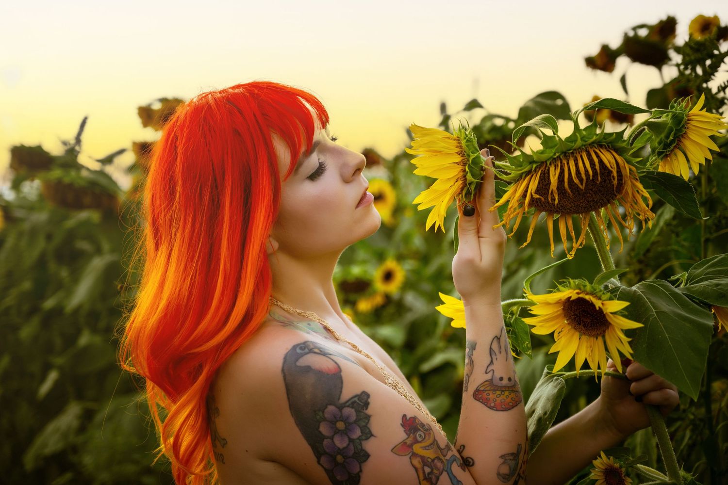 Woman with vibrant red hair and tattoos touching sunflowers in a field at golden hour.