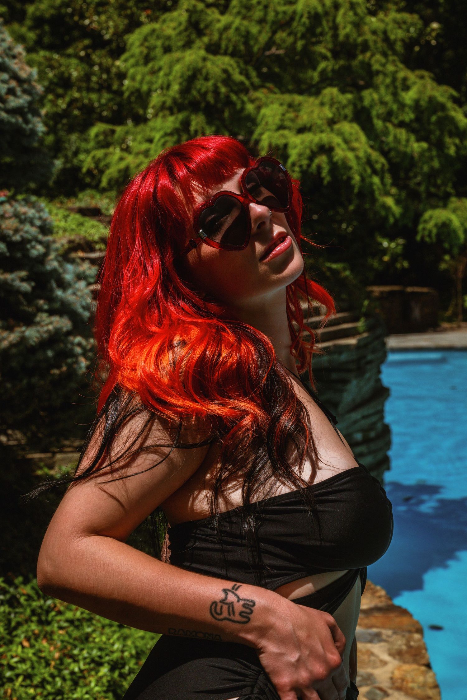 Woman with vivid red hair and sunglasses posing in black outfit near a pool.